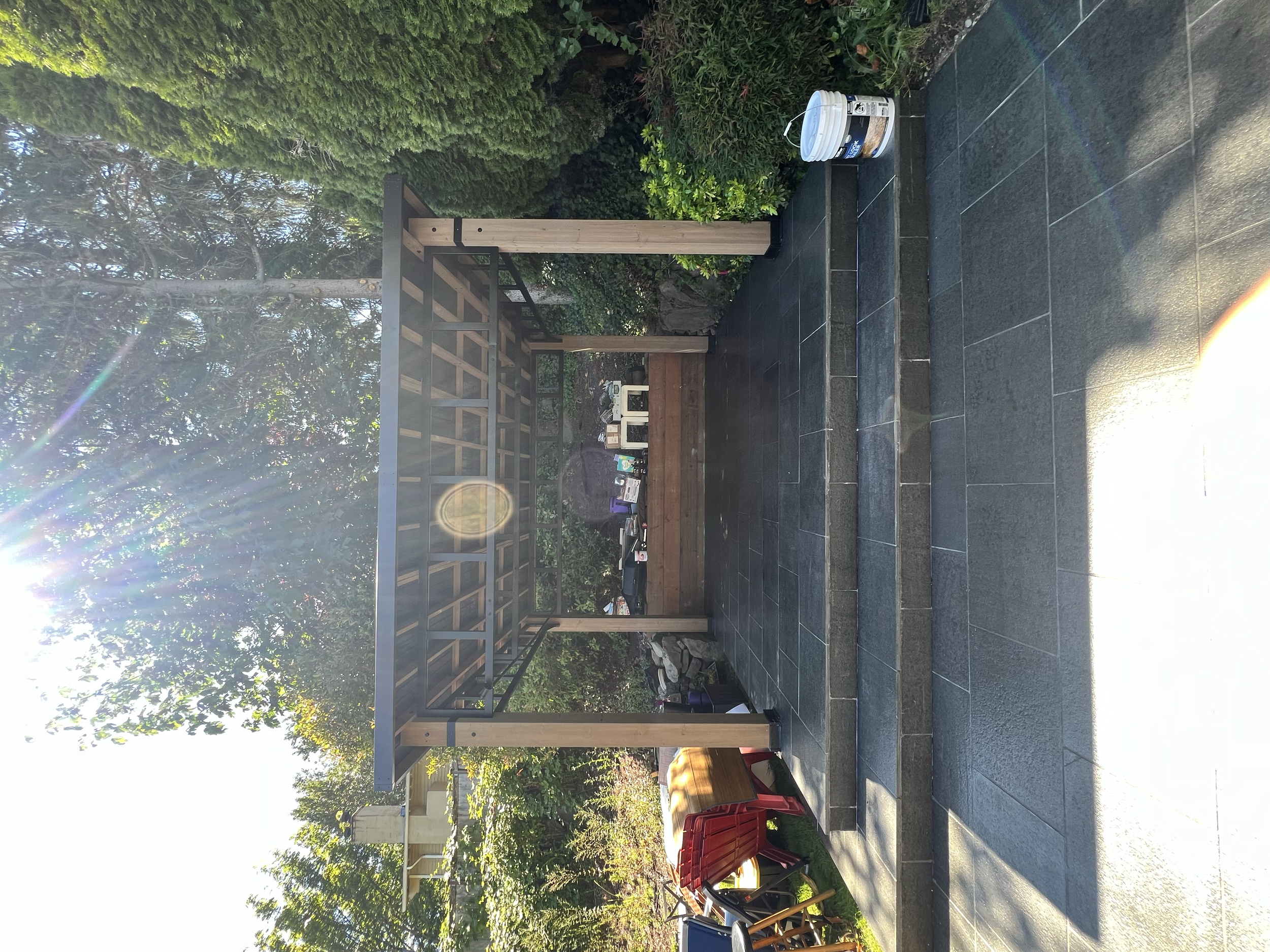 A backyard patio with black tiled flooring and a black metal and wood canopy. There are trees and greenery in the background and some gardening or construction supplies on the ground.