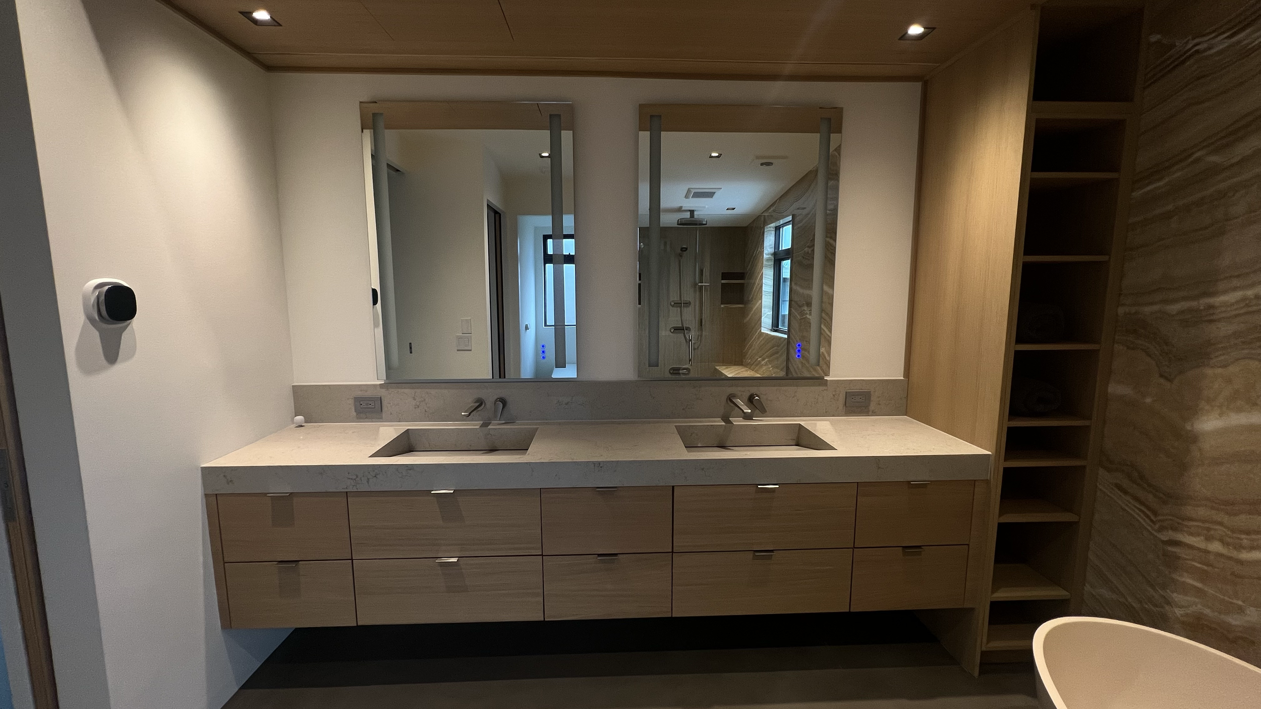Modern bathroom with double sinks, wooden cabinets, a large mirror, and a shower area with a window visible in the reflection.