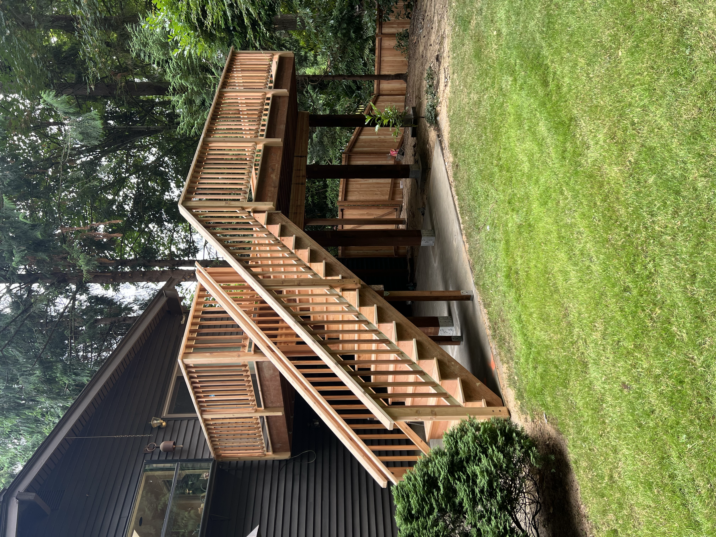 Wooden deck with stair railing attached to a black house with a window and outdoor light in a grassy backyard with trees and bushes.