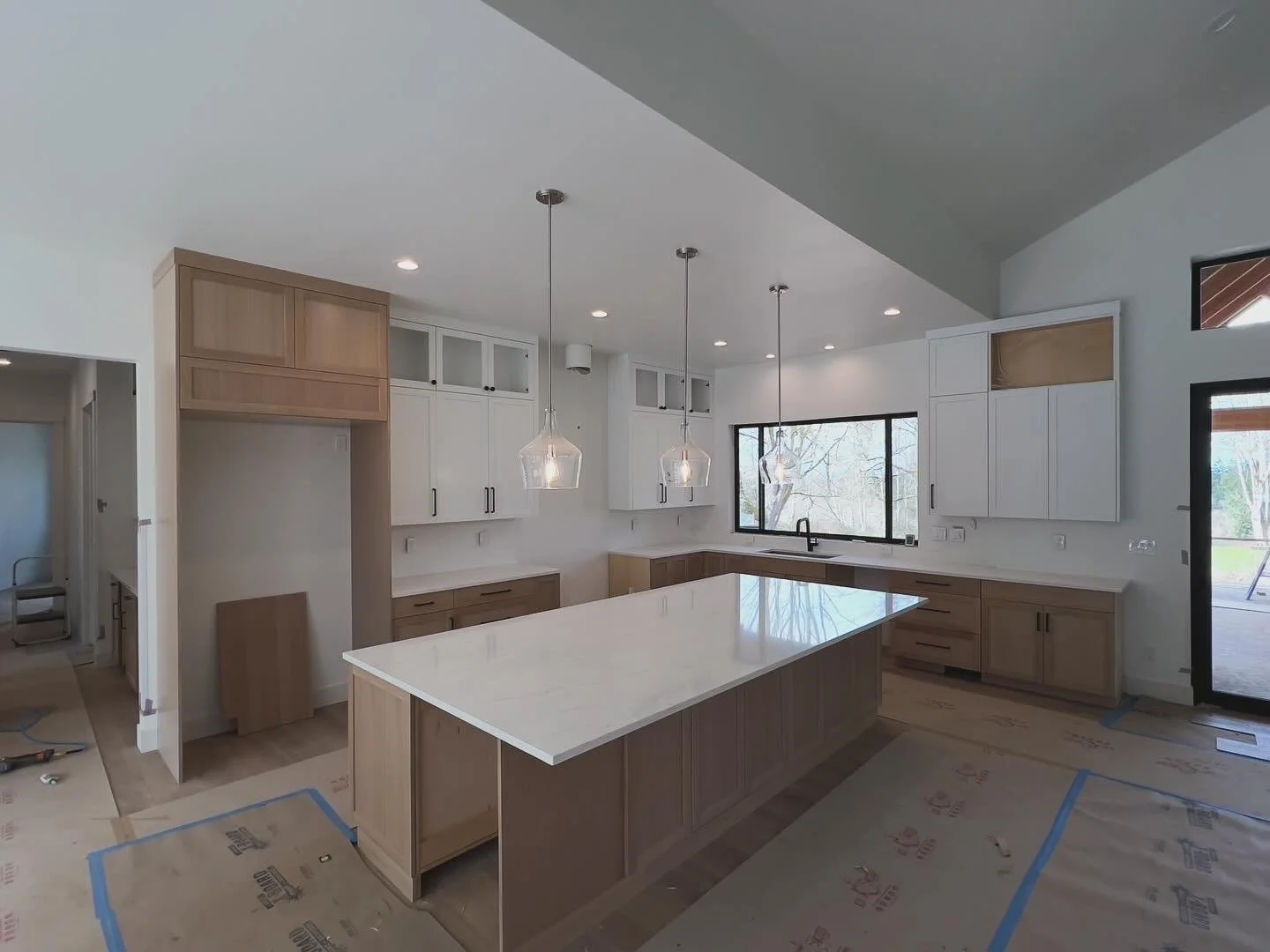 Modern kitchen under construction with white and light wood cabinetry, a large island with a white marble countertop, three pendant lights above, and a large window with a view of trees outside.