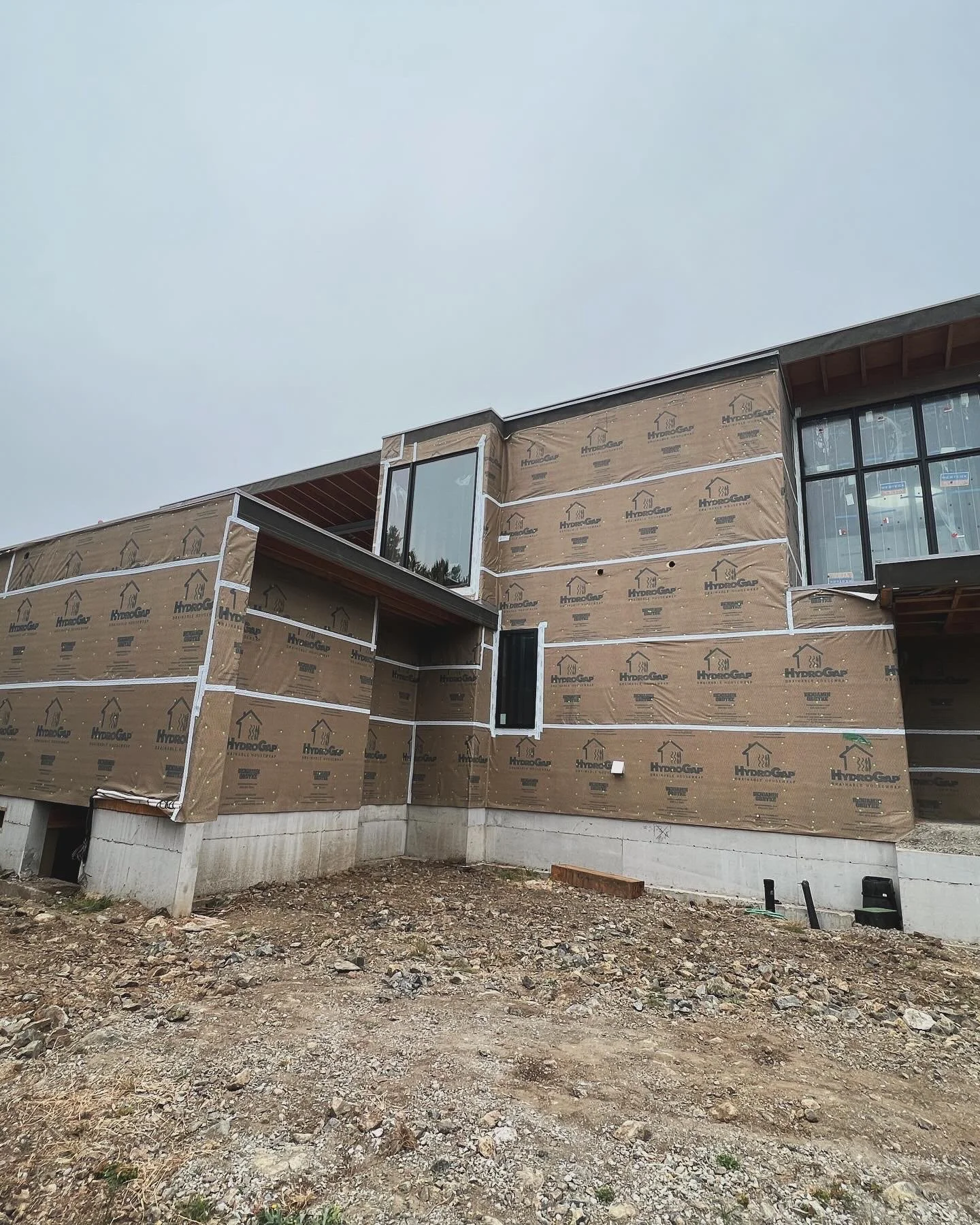 A building under construction with exterior walls covered in brown weather-resistant sheathing and visible window frames, on a dirt lot with an overcast sky.