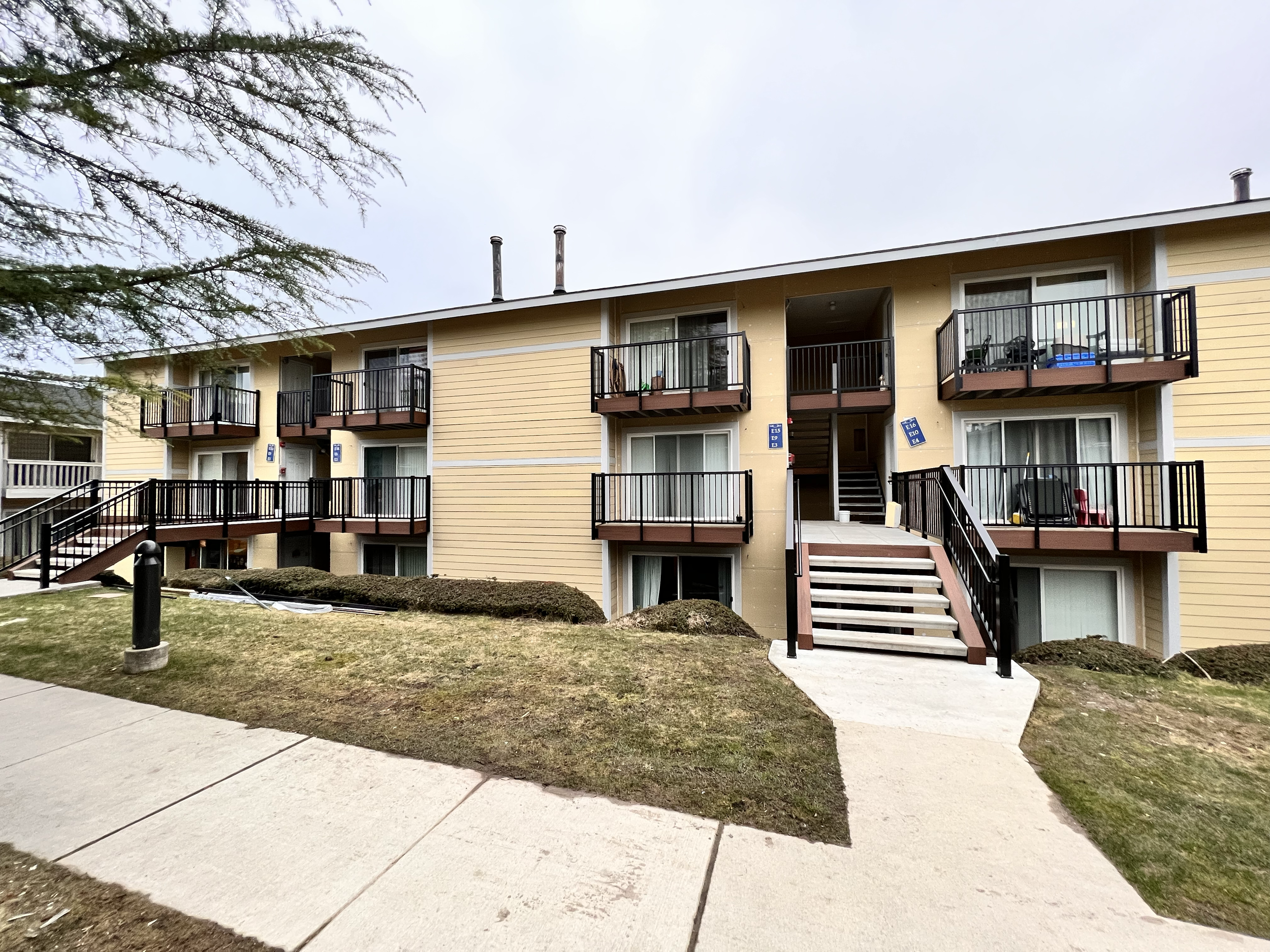 Multi-story apartment building with balconies, stairs, and a sidewalk in front, on a cloudy day.
