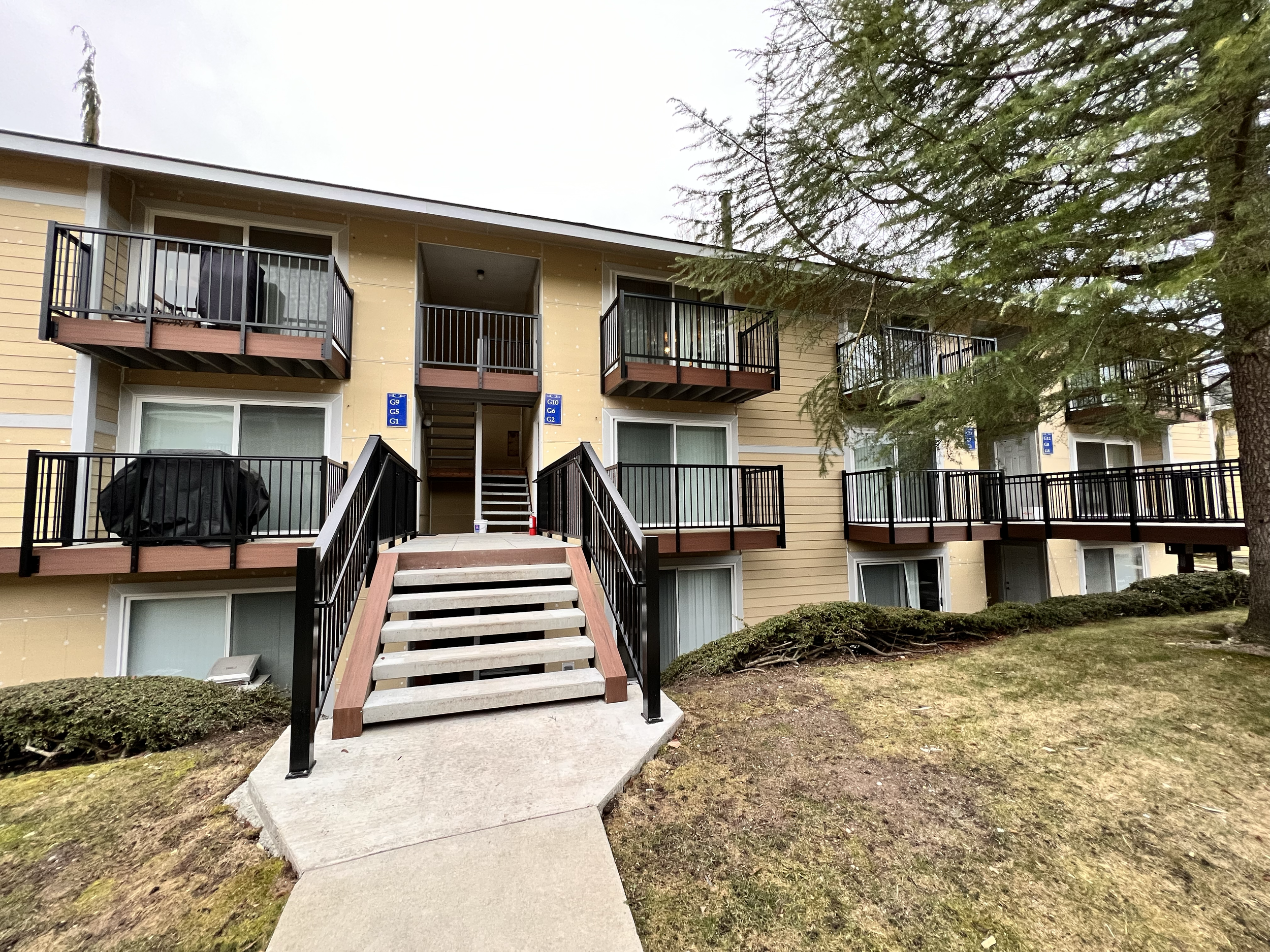 Exterior view of a beige apartment building with balconies and stairs leading to the second floor, surrounded by grass, bushes, and a large tree.