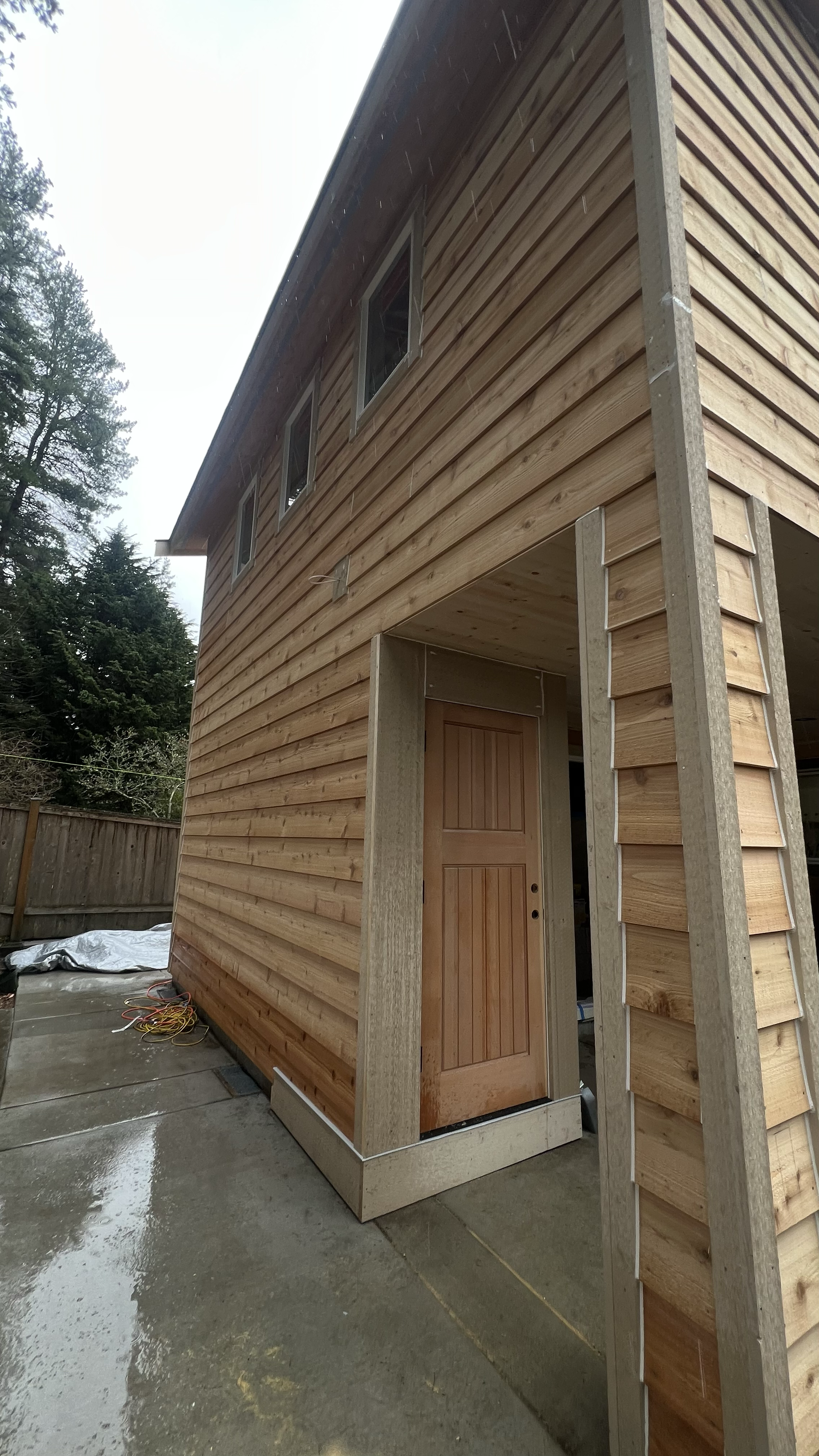 Construction site of a two-story house with wooden siding, multiple windows, and a doorframe without a door, with wet concrete and construction materials on the ground.