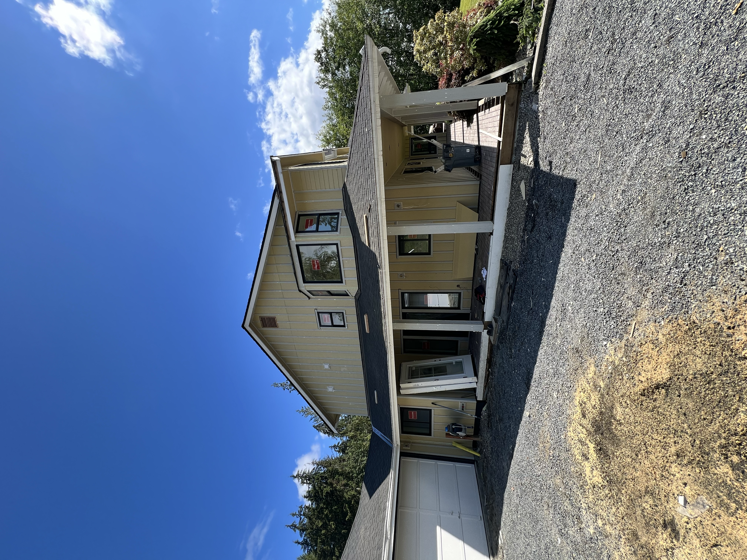 A two-story beige house under construction with a black shingle roof, a white garage door, and a front porch with a white railing. There is gravel in the driveway and some construction tools on the porch. The sky is clear with a few clouds, and there