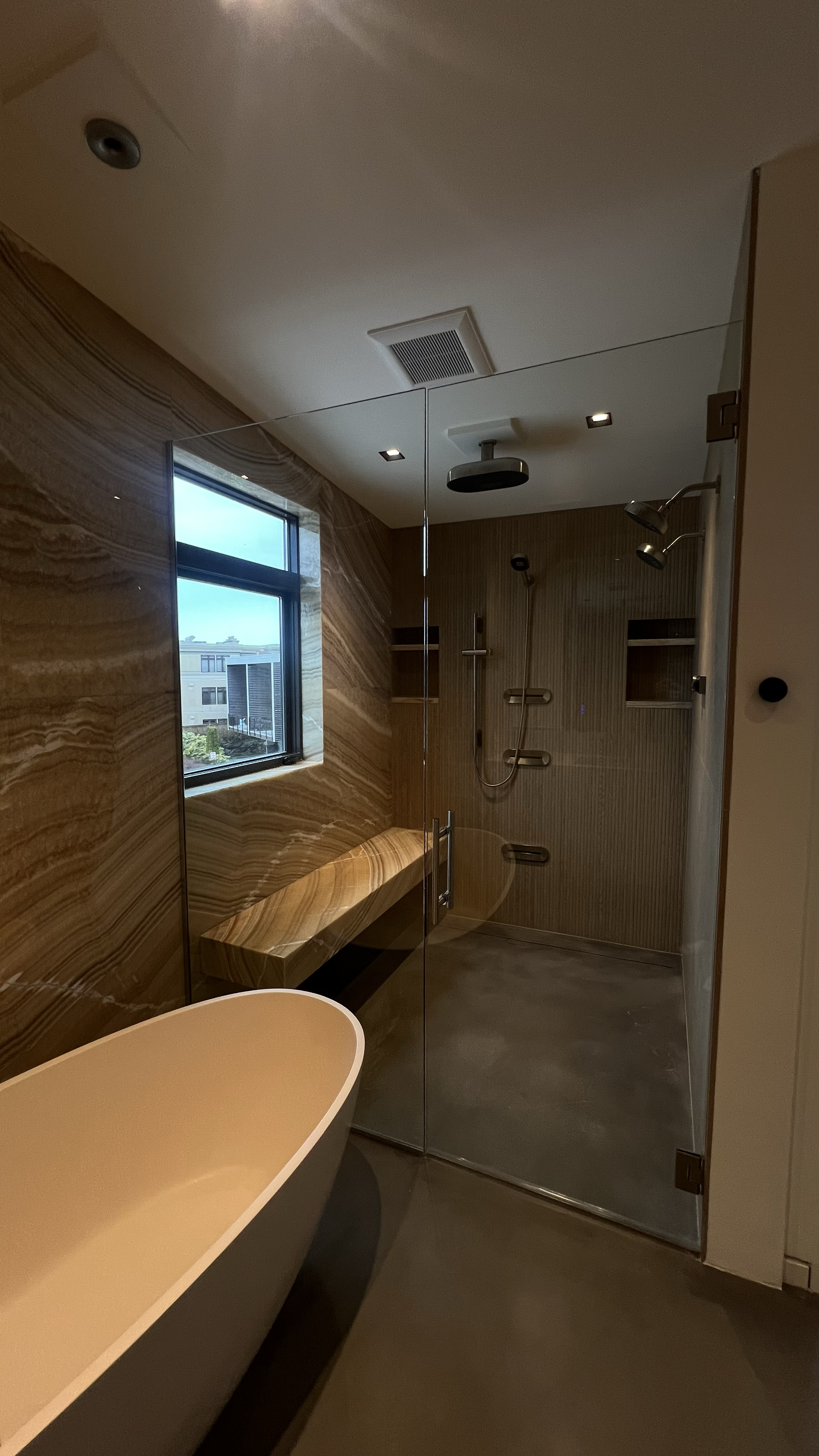 Modern bathroom with a freestanding bathtub, a wooden bench, and a walk-in shower with multiple showerheads, a wooden wall, and a window.