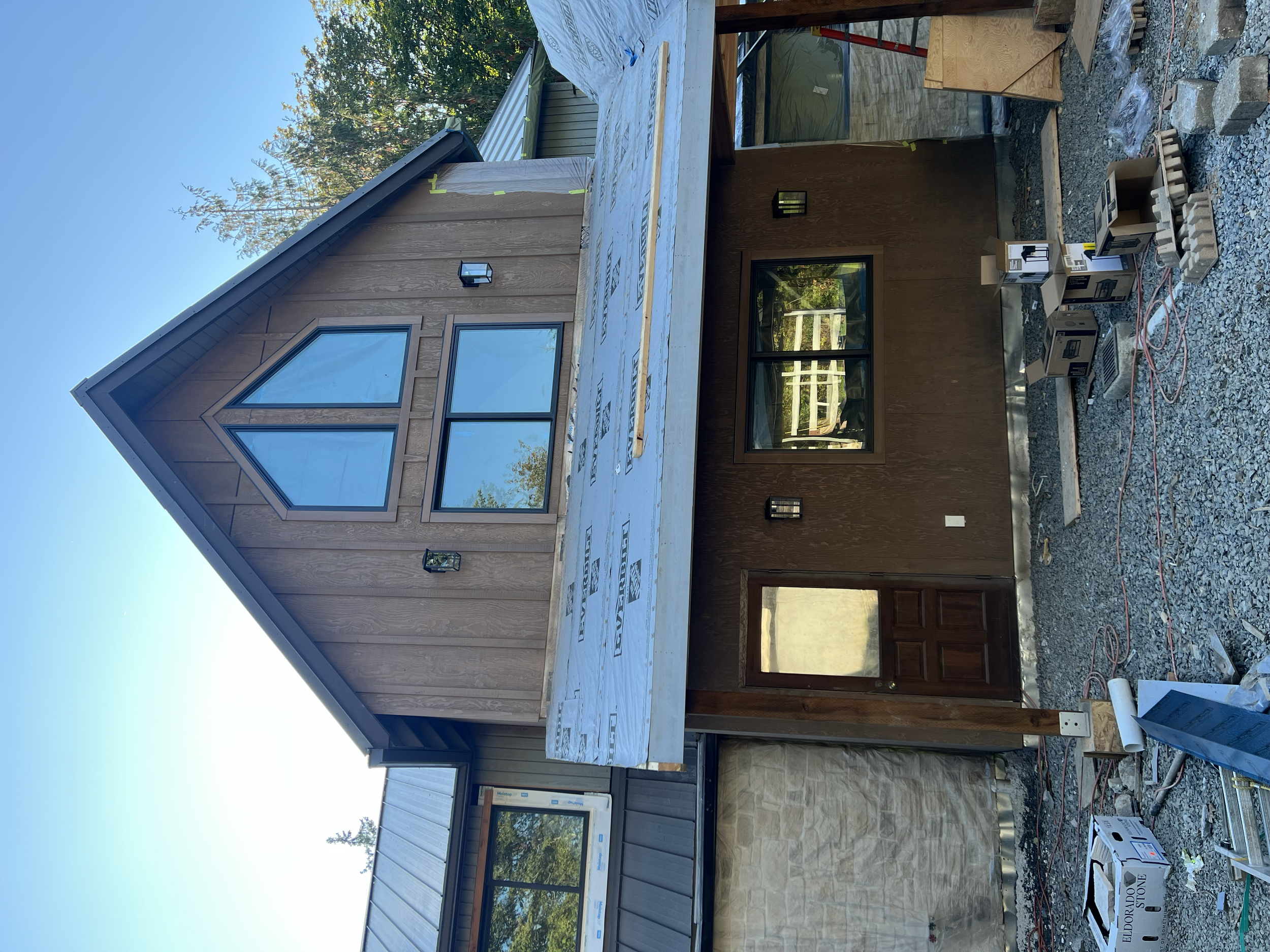 A two-story house under construction with wood siding on the upper part and brick on the lower part. The roof has a steep pitch with a large diamond-shaped window and a smaller rectangular window below it. There is a window and a door on the lower le