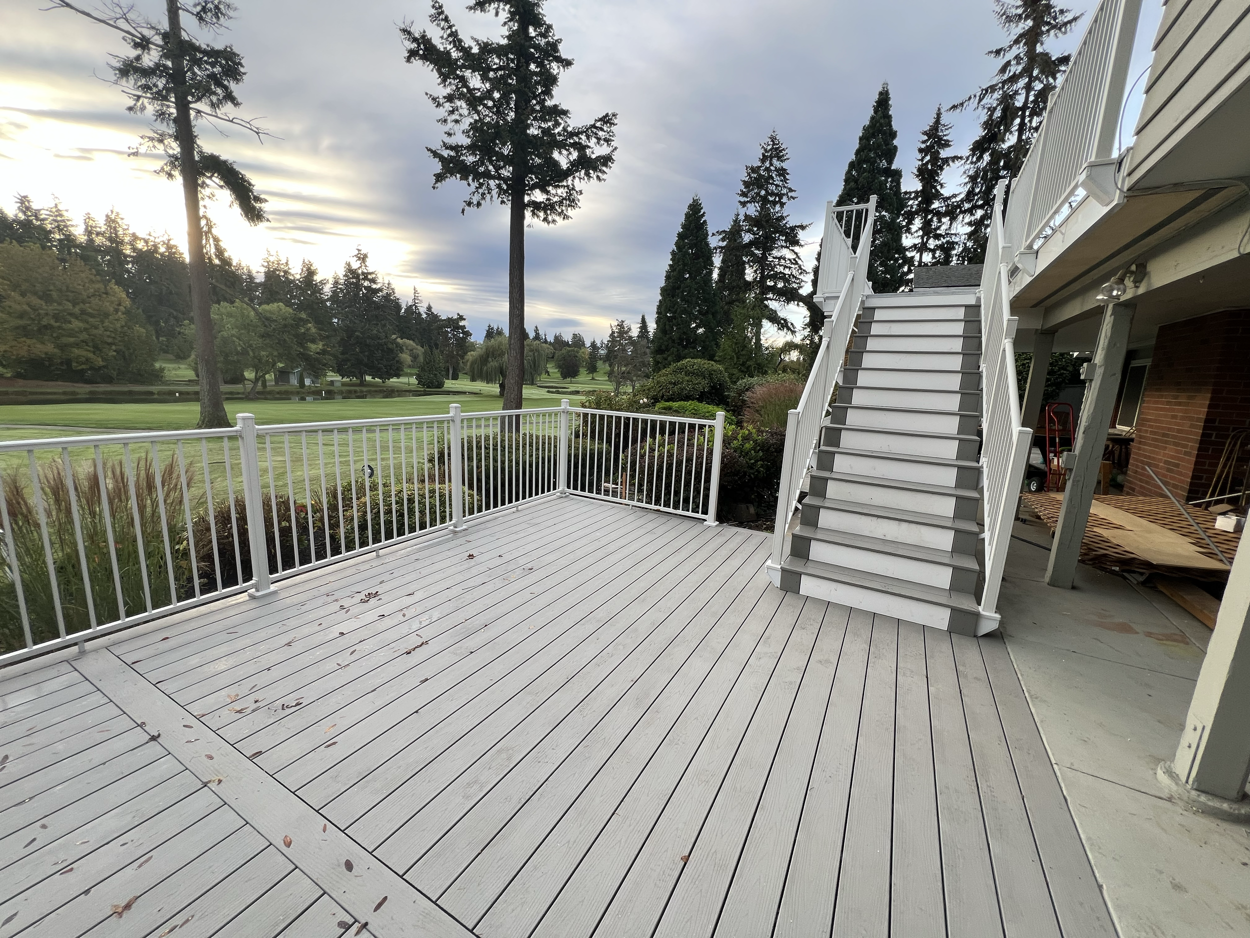 Empty outdoor deck with white railings and stairs leading to an upper level, overlooking a golf course with trees, grass, and a cloudy sky.