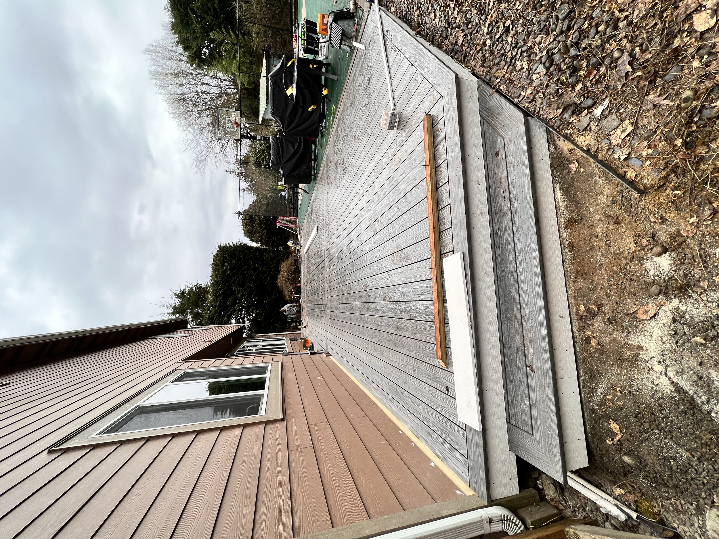 A newly built wooden deck attached to a house with brown siding, showing part of a window, with surrounding gravel and dirt, and construction materials and tools on the deck.