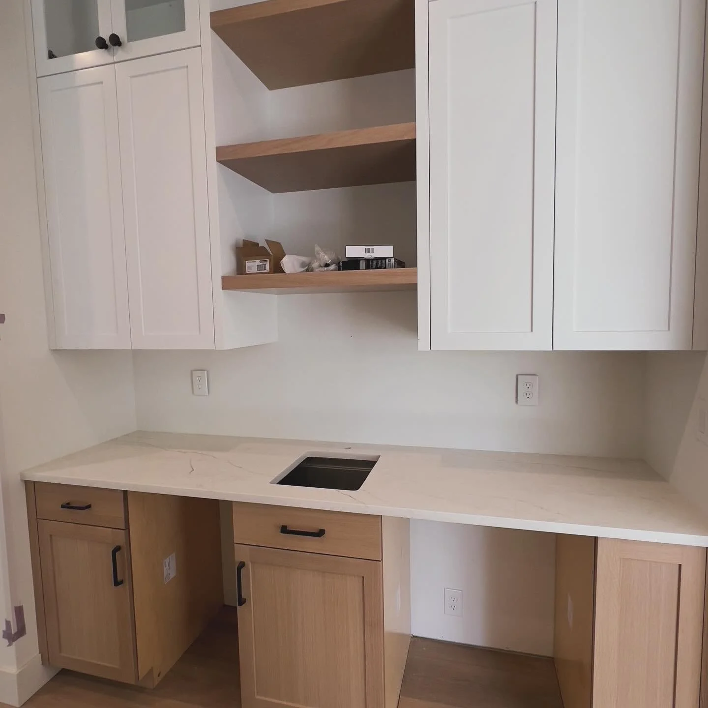Kitchen cabinets with upper white doors, open wooden shelves, lower light wood cabinets, and a white marble countertop with a cutout for a sink.