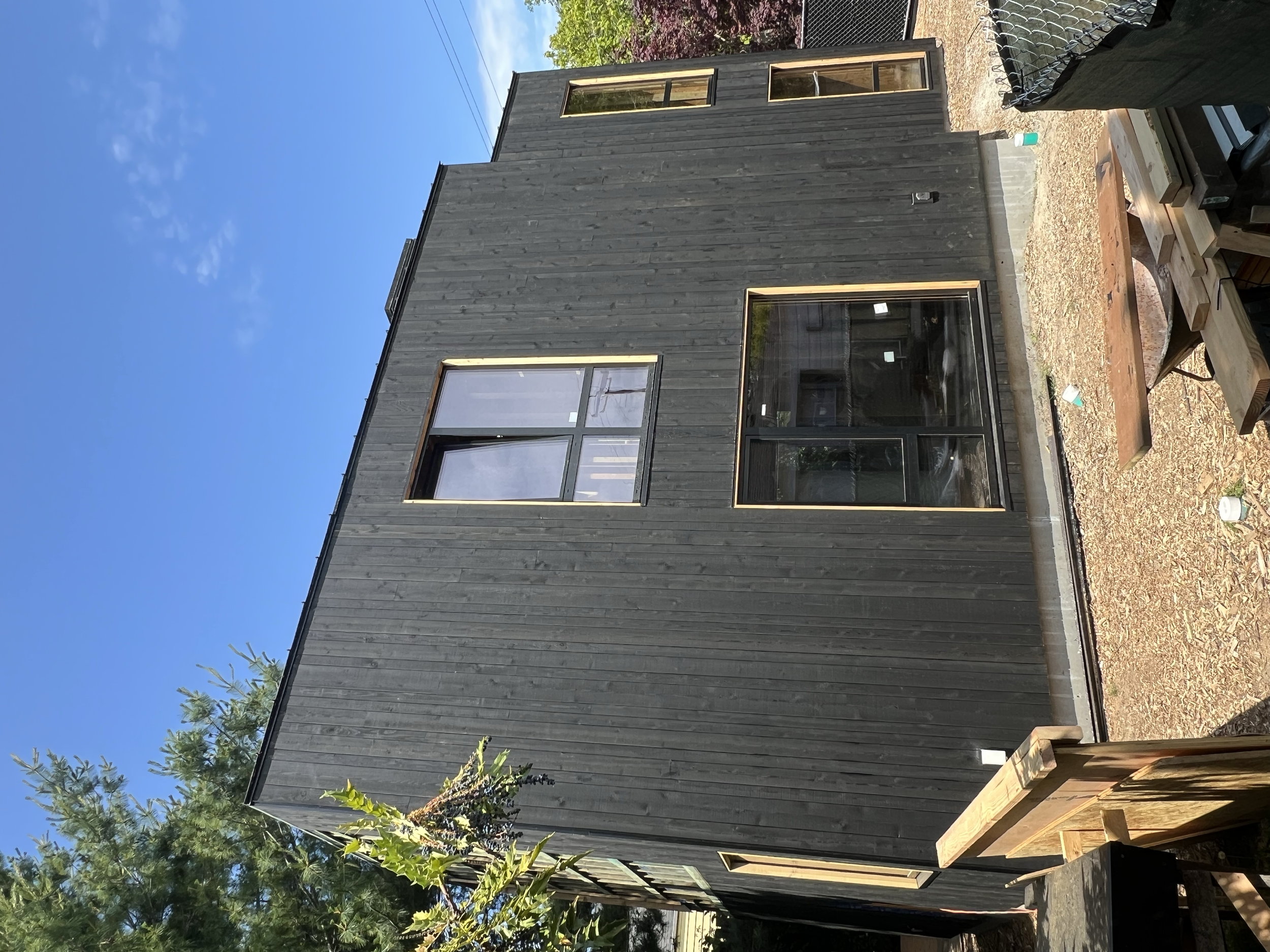 An exterior view of a two-story black wooden house with large windows, surrounded by trees and a gravel yard, under a clear blue sky.