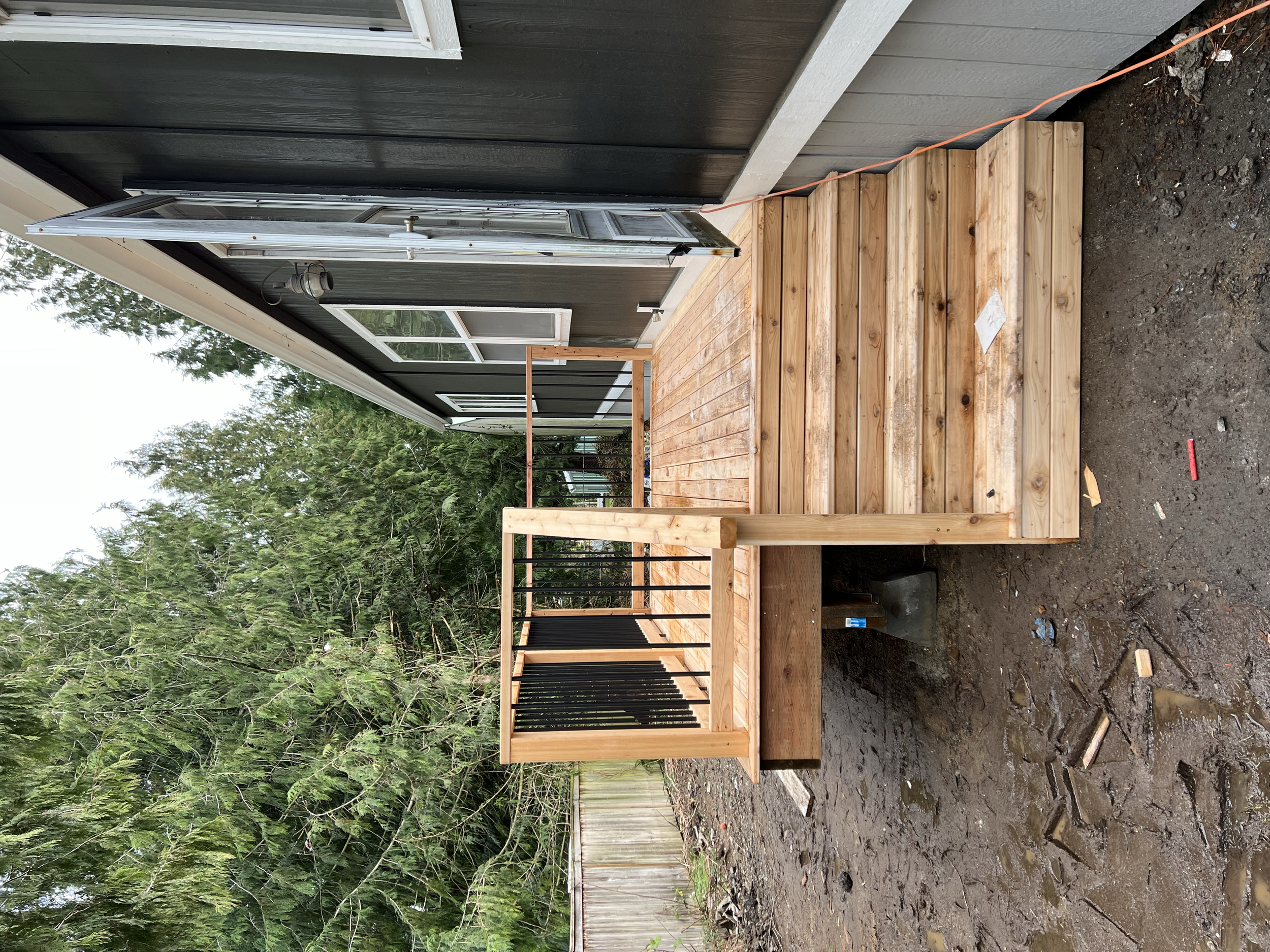 A wooden deck under construction next to a house with dark green siding and a window, with trees and a wooden fence in the background.