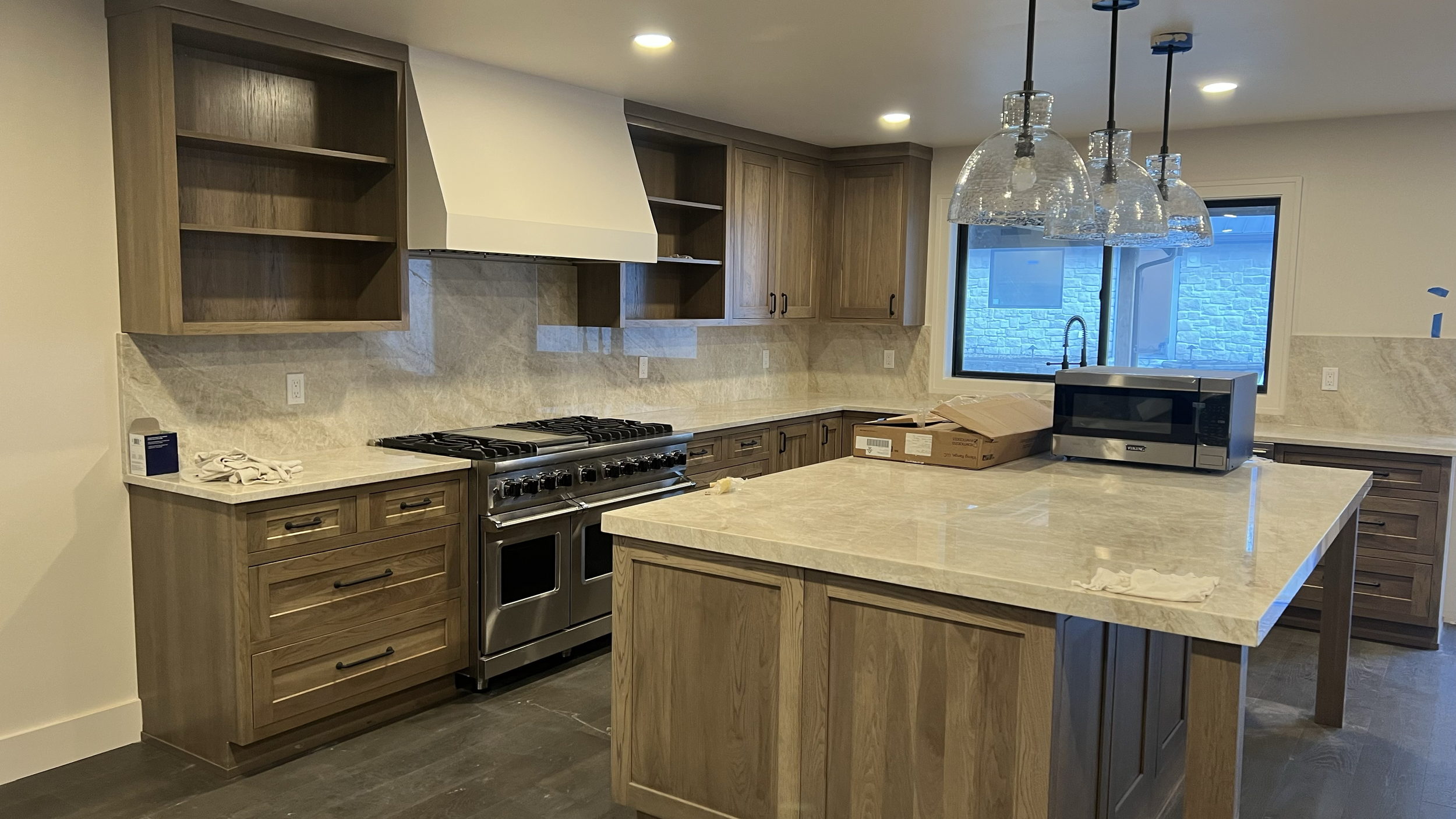 Kitchen with wooden cabinets, a stainless steel gas stove, countertop, kitchen island, microwave, and a window showing a stone wall outside.