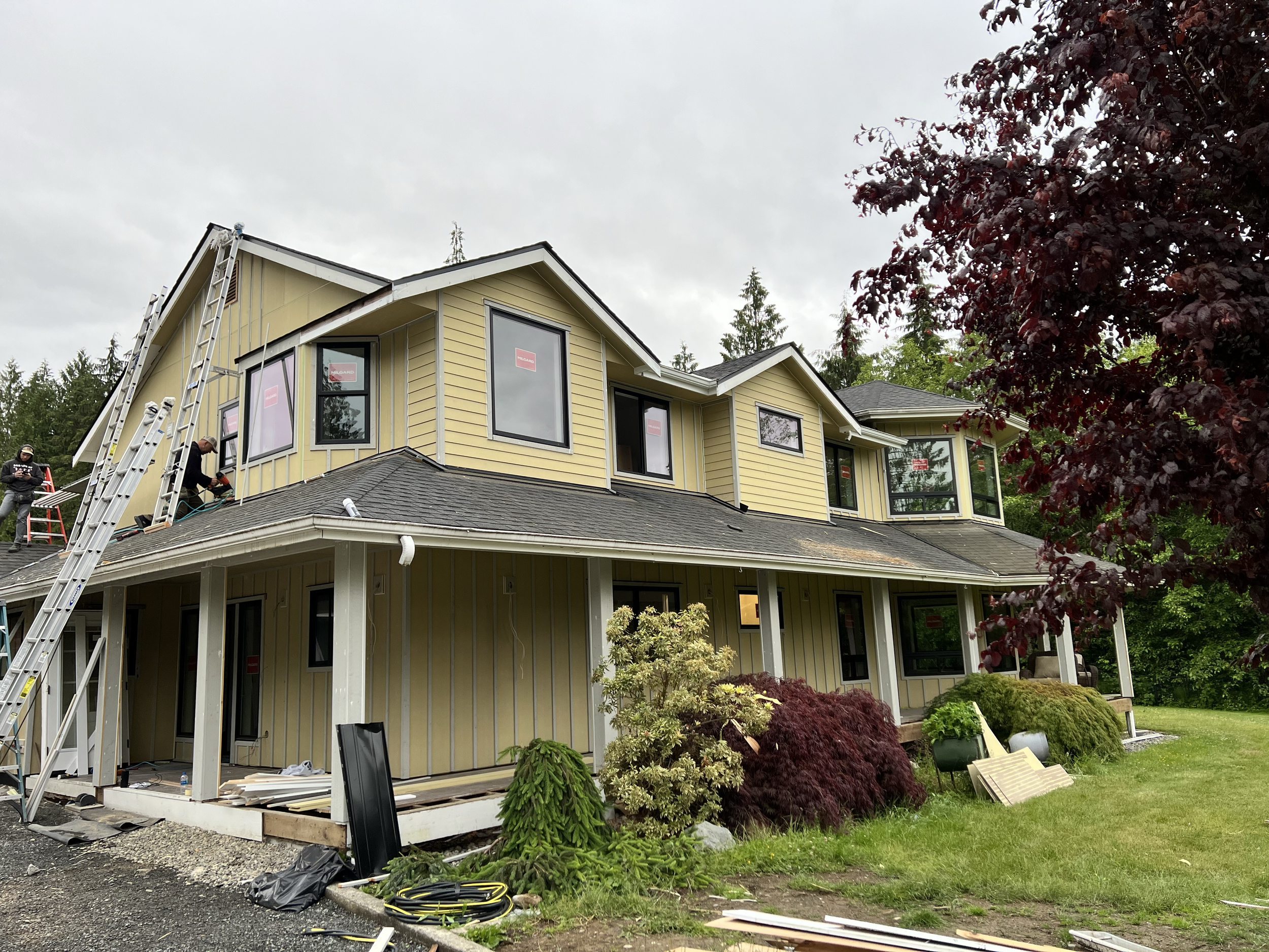A large, two-story yellow house under construction with workers on the roof and ladders. The house has multiple windows, some with red signs, and is surrounded by greenery, including trees and shrubbery.
