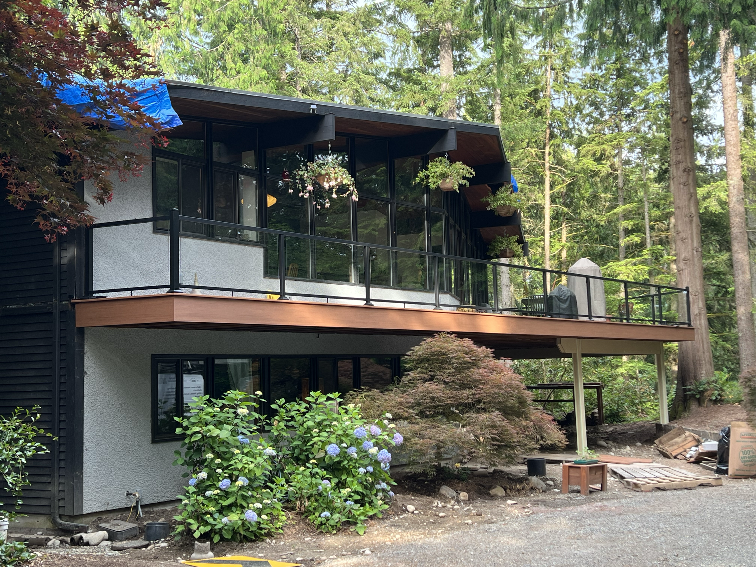 Modern two-story house with large glass windows and a spacious balcony, surrounded by tall trees and lush greenery.
