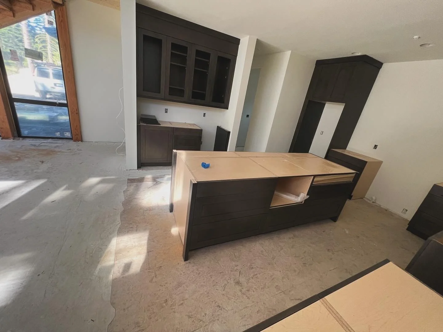 Under construction kitchen with dark wood cabinets and an island, unfinished flooring, and a sliding glass door bringing in natural light.