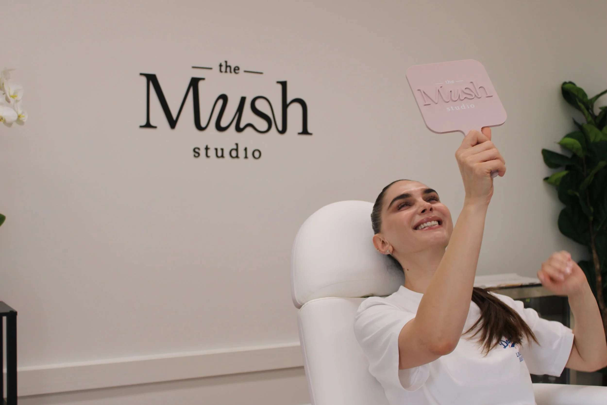 A woman smiling and holding a pink sign with the words 'the Mush studio' inside, sitting in a lounge chair at The Mush studio, with the studio's logo on the wall behind her.