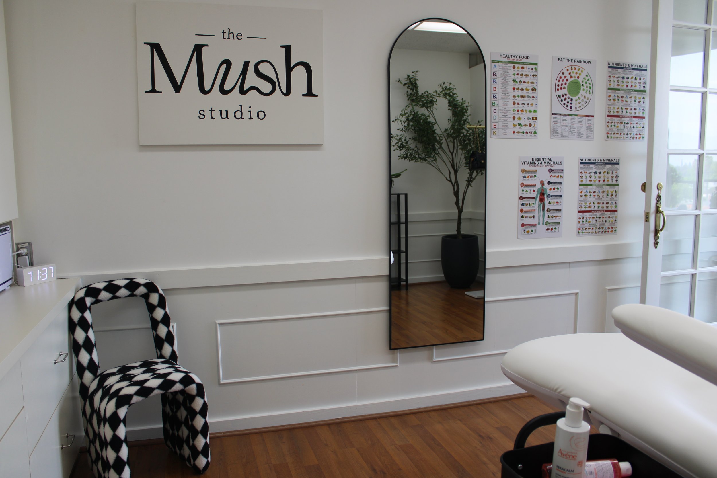 Interior of a wellness or treatment center with a sign that reads 'the MUSH studio'. There are health and nutrition posters on the wall, a full-length mirror, a salon chair with a black and white checkered pattern, a white treatment table with bottles of skincare products, and a door with glass panels leading outside.