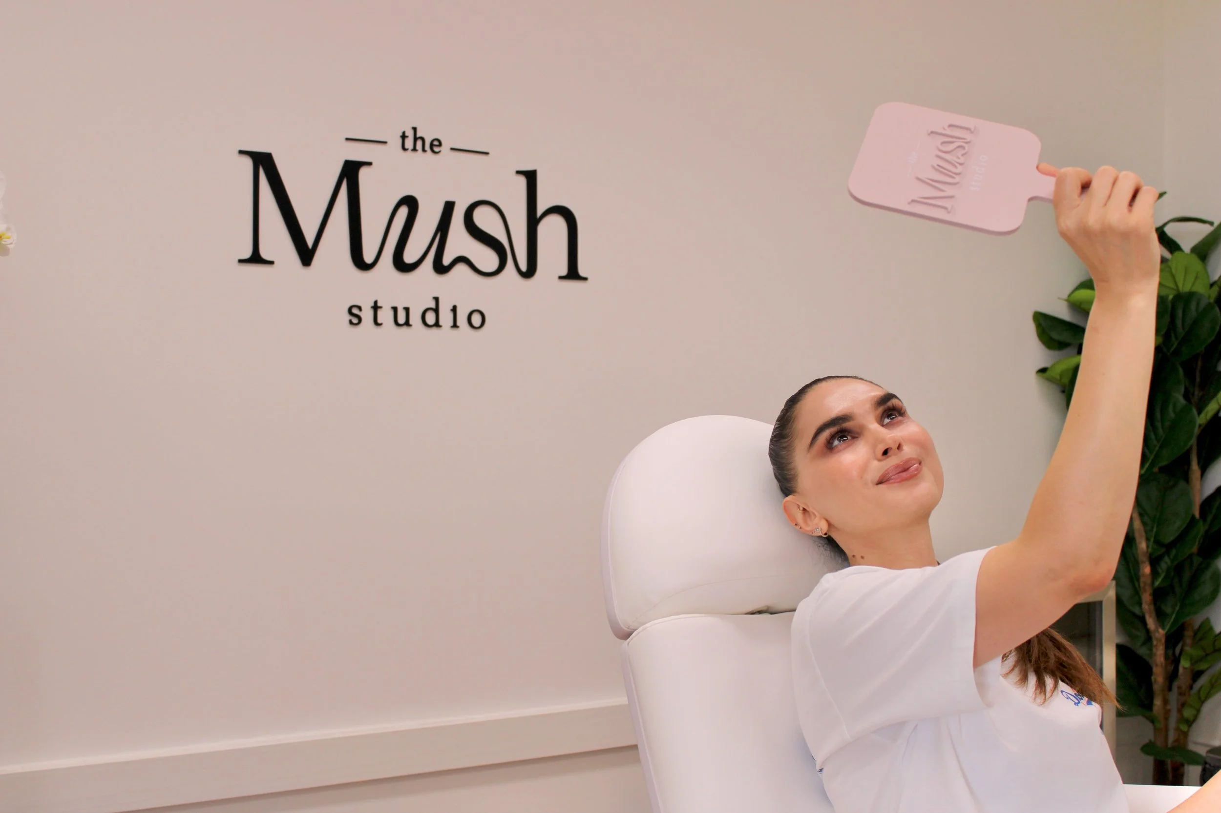 A woman relaxing in a spa chair holding a pink paddle with the logo 'Mask' printed on it, in front of a wall with the text "the Mush studio."
