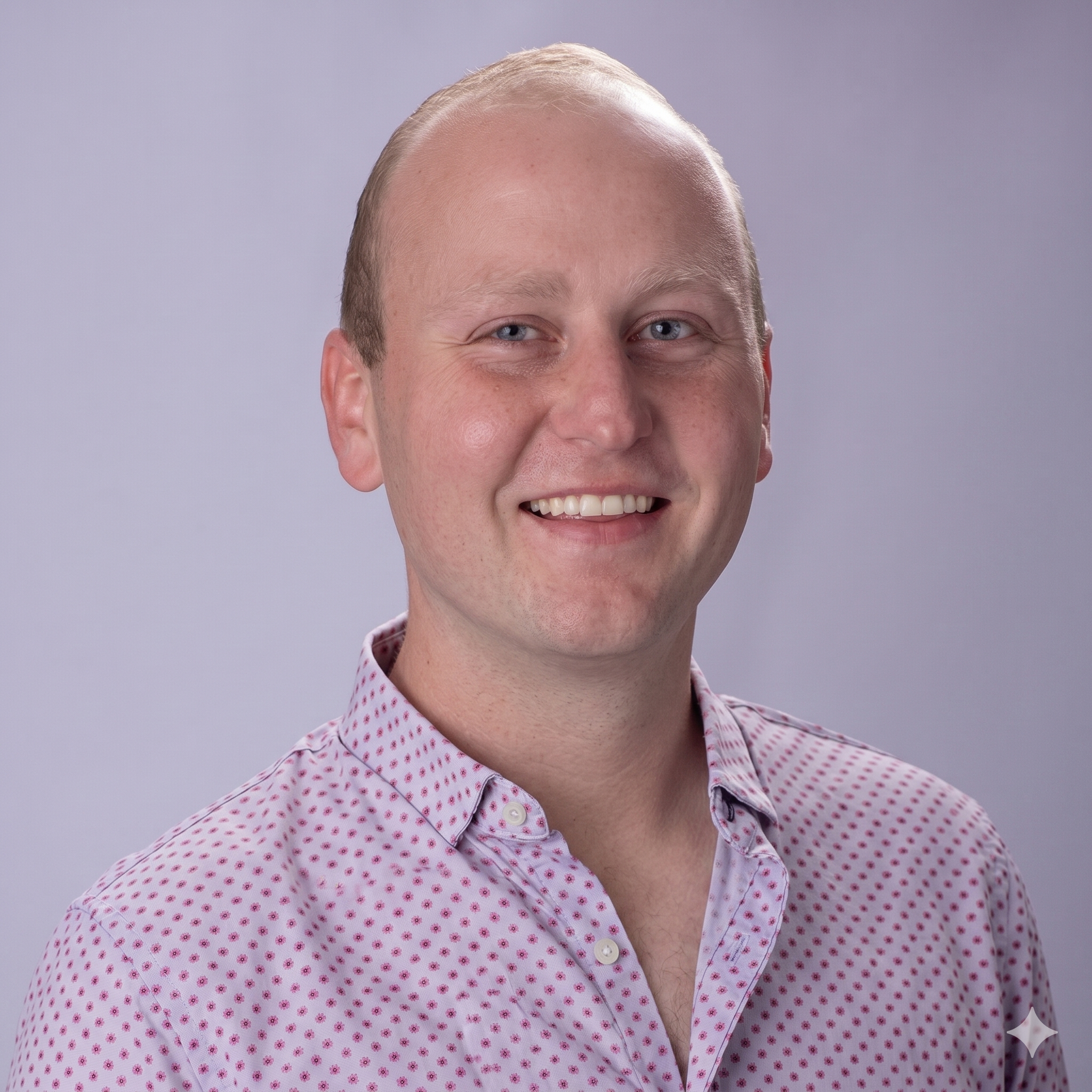 A young man with short blonde hair, wearing a pink and white polka dot button-up shirt, smiling against a light gray background.