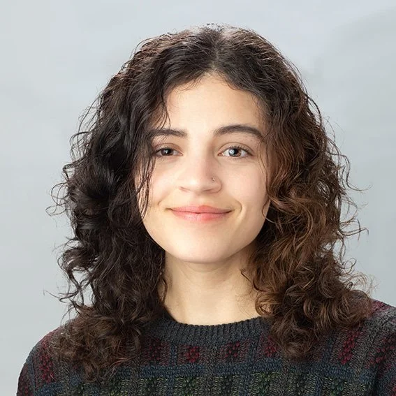 A young woman with curly brown hair, smiling, wearing a dark sweater with a colorful pattern, against a plain light gray background.