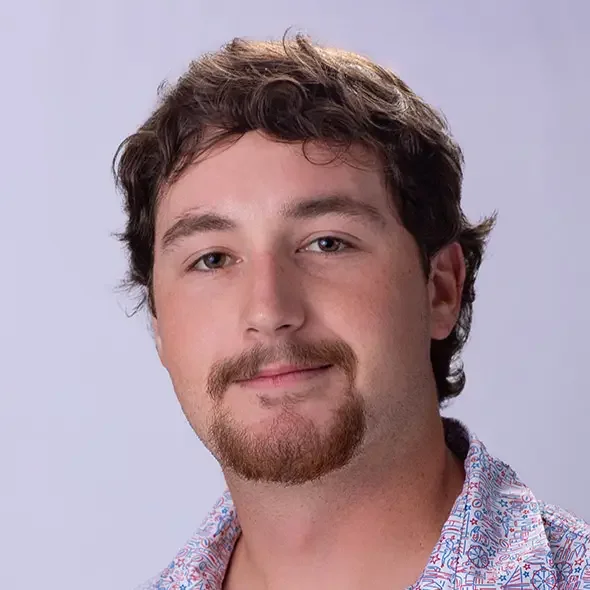 A man with curly brown hair, blue eyes, and light skin, wearing a patterned shirt, looking at the camera against a plain background.