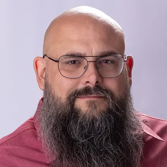 A middle-aged man with a bald head, glasses, and a full beard wearing a maroon shirt against a light gray background.