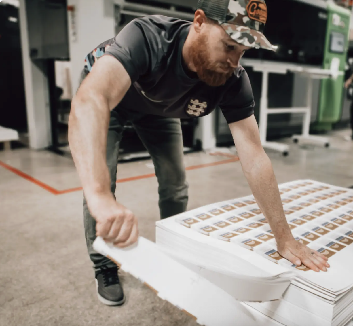 A man in a camouflage hat and dark shirt leaning over a stack of printed posters or flyers, pulling one away in an indoor office or workshop setting.