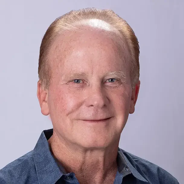 Headshot of an older man with light skin, light hair, and blue eyes, wearing a blue collared shirt against a plain background.