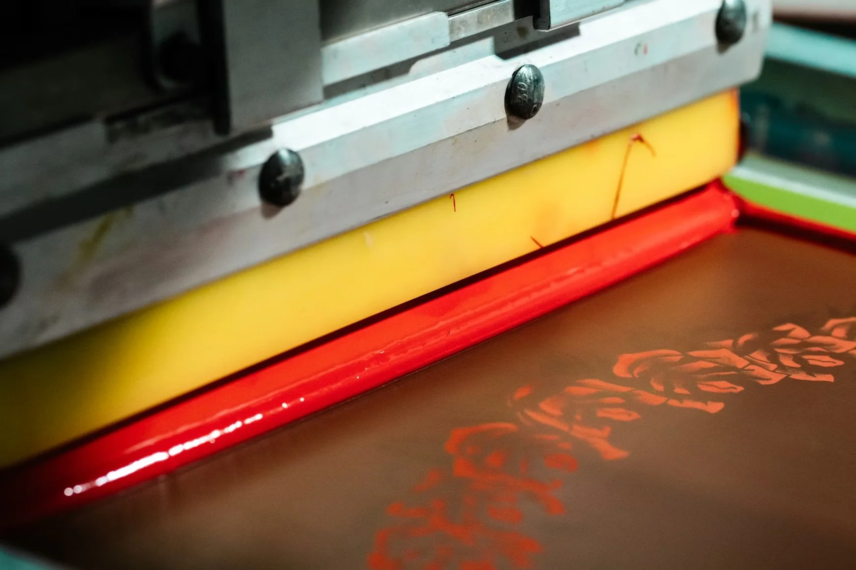 Close-up of a screen printing machine with a red, yellow, and green color palette and a partially printed orange design on a dark surface.