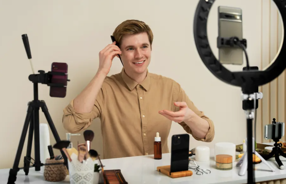 Young man recording a video or live stream in a home setup with ring light, phone, and makeup products on the table.