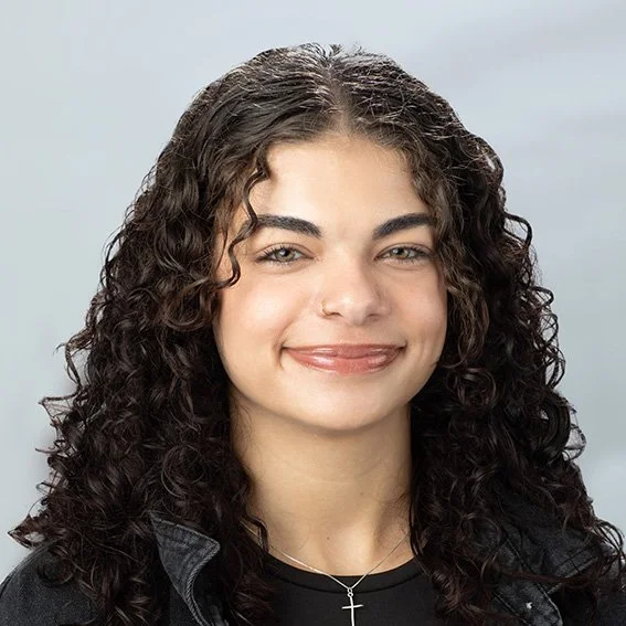 A young woman with curly dark hair smiling at the camera, wearing a black top and a silver cross necklace.