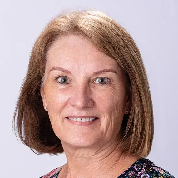 A woman with shoulder-length light brown hair, wearing a floral top, smiling against a light gray background.
