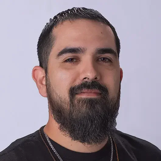 Close-up photo of a man with dark hair, a full beard, and parted hair, wearing a black shirt and silver necklaces, against a light gray background.