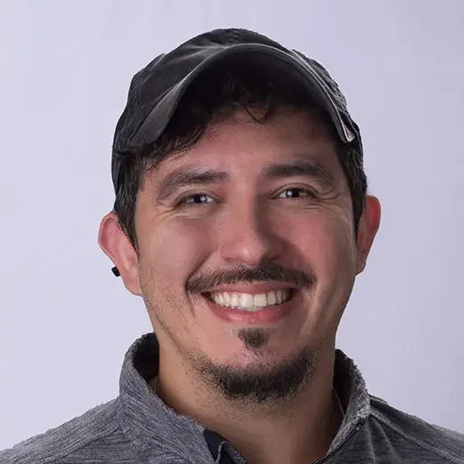 A smiling man wearing a dark baseball cap and a gray zip-up jacket against a light gray background.