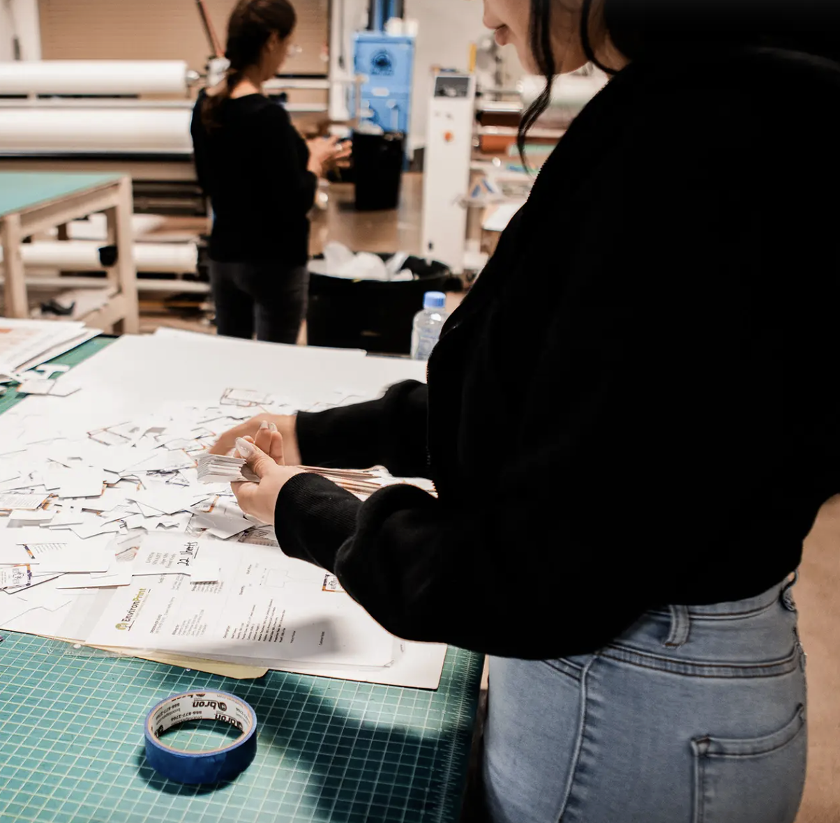 Person with dark hair wearing a black jacket and light blue jeans working on a project at a workshop table with papers, tape, and supplies, while another person works in the background in a creative workshop or studio.