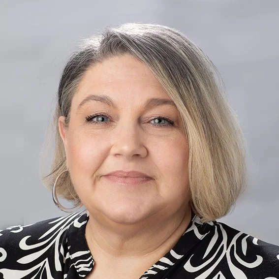 A woman with shoulder-length blonde hair, wearing a black and white patterned top and hoop earrings, smiling at the camera against a plain gray background.