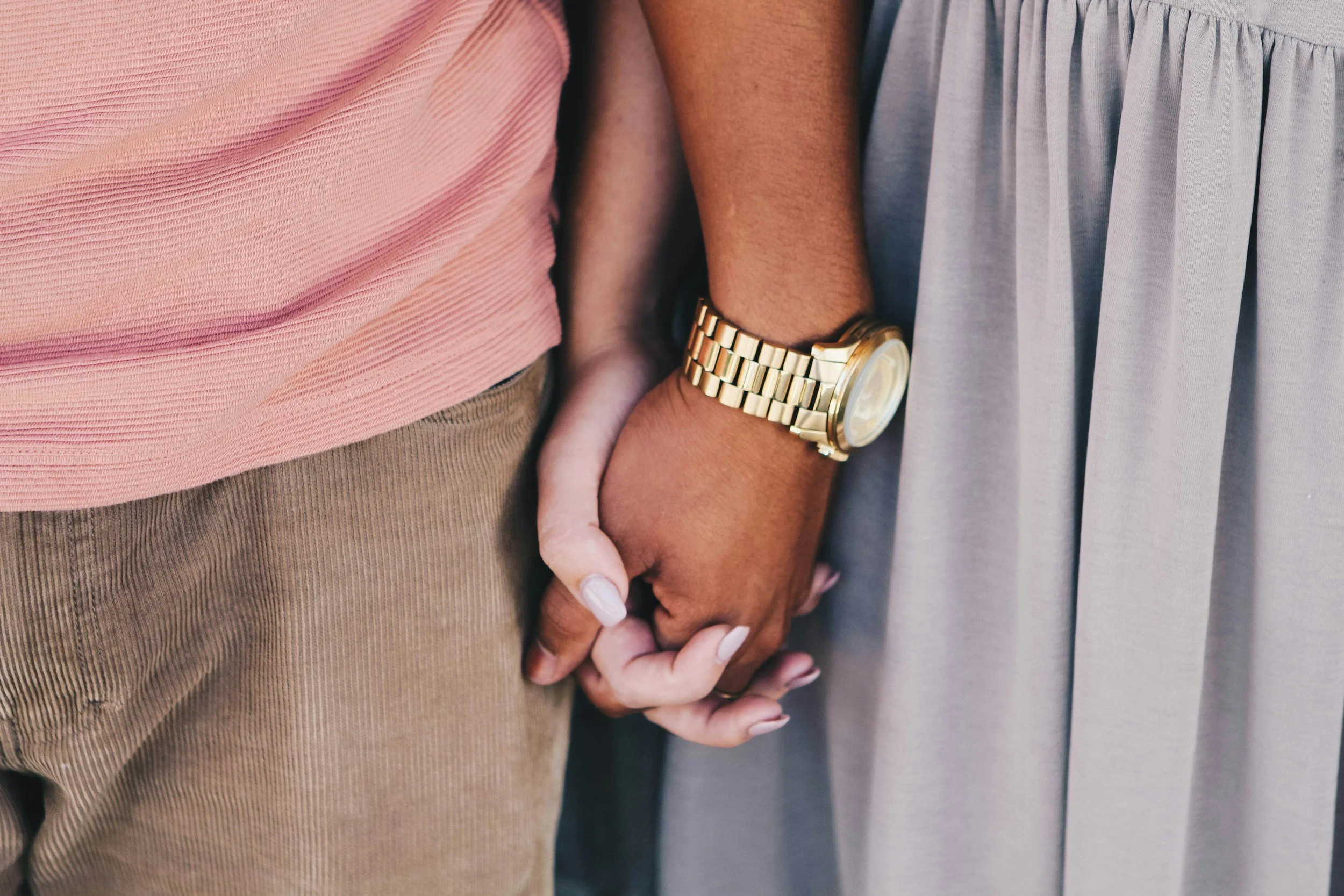 Close-up of two people holding hands, one wearing a pink shirt and beige pants, the other wearing a light gray dress and a gold watch.