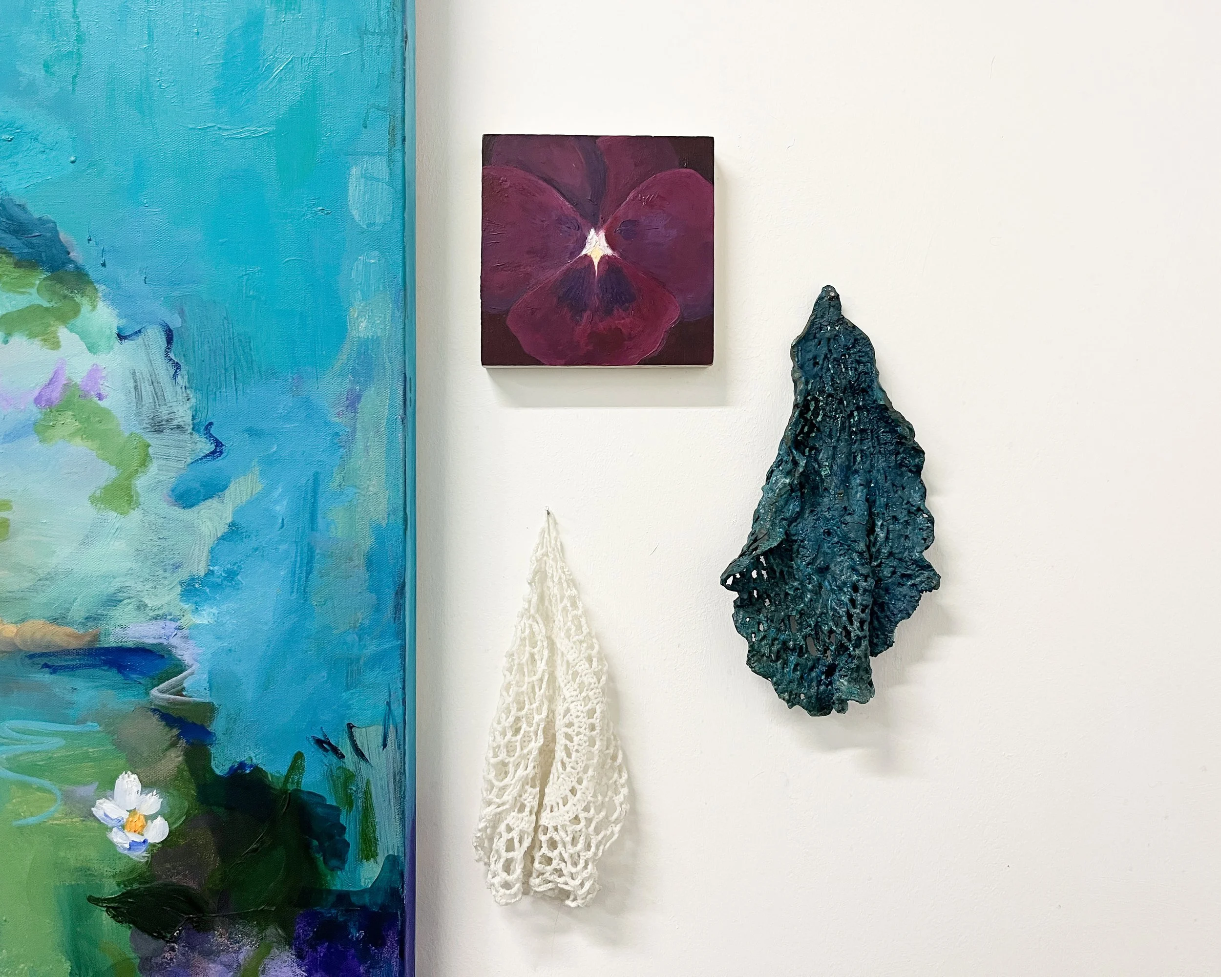 Art displayed on a white wall with a colorful abstract painting, a small purple floral painting, a white crocheted piece, and a dark textured sculpture.