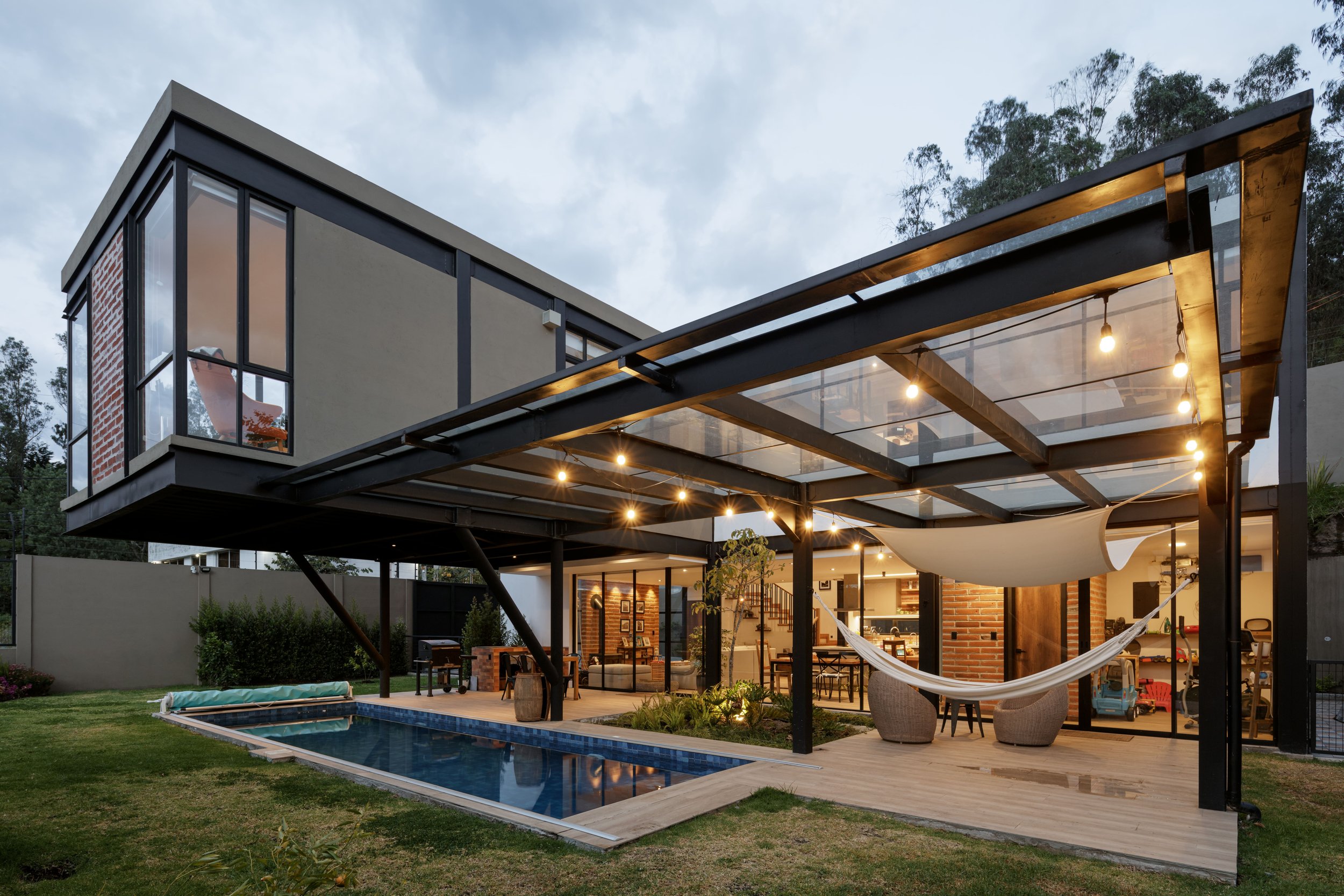 Casa moderna con balcón de vidrio, jardín con piscina y área de descanso con hamacas y sillones, iluminada en la noche.