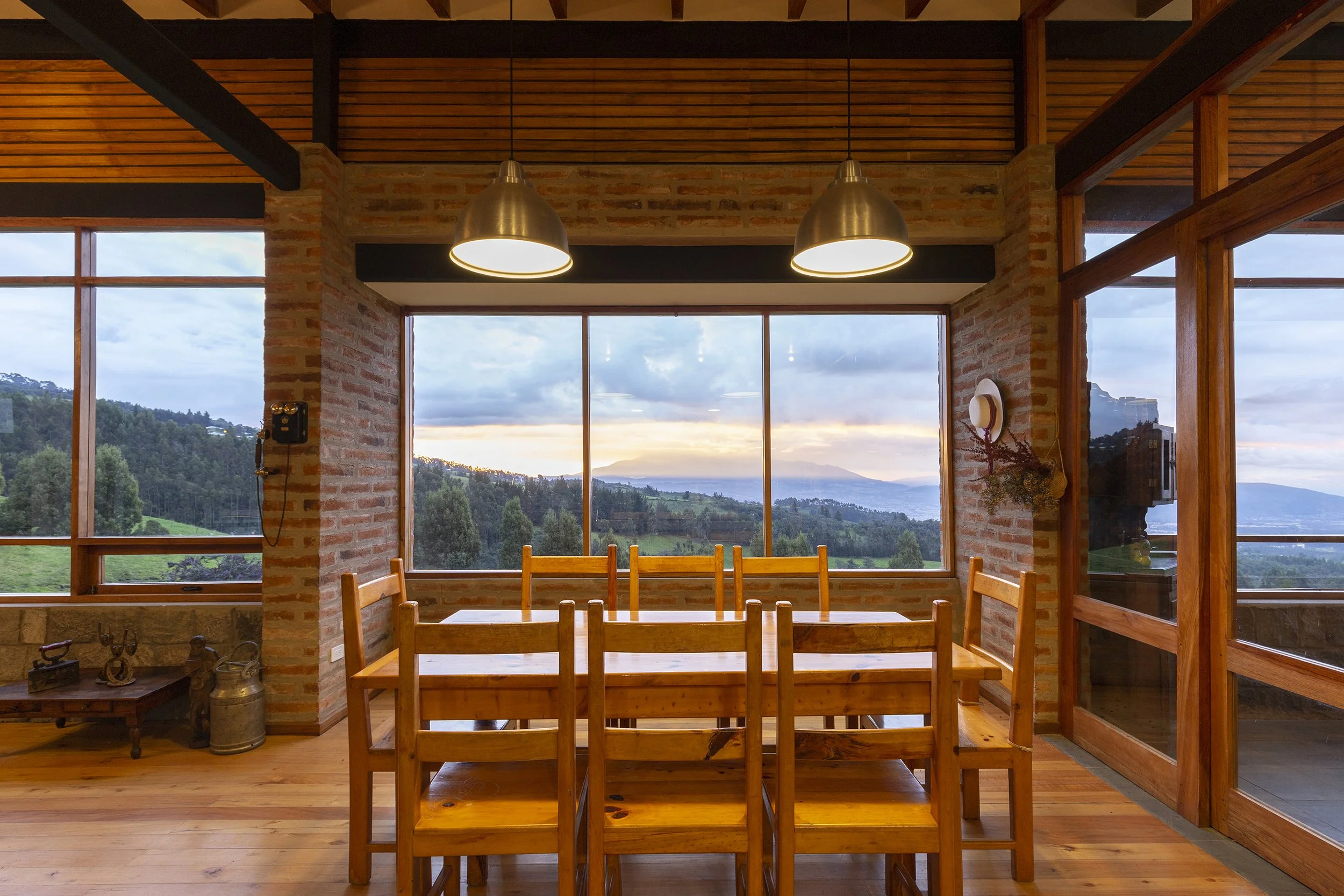 Interiores de una casa con paredes de ladrillo, grandes ventanas que muestran un paisaje de montañas y árboles, y una mesa de madera con sillas, iluminadas por lámparas colgantes de metal.