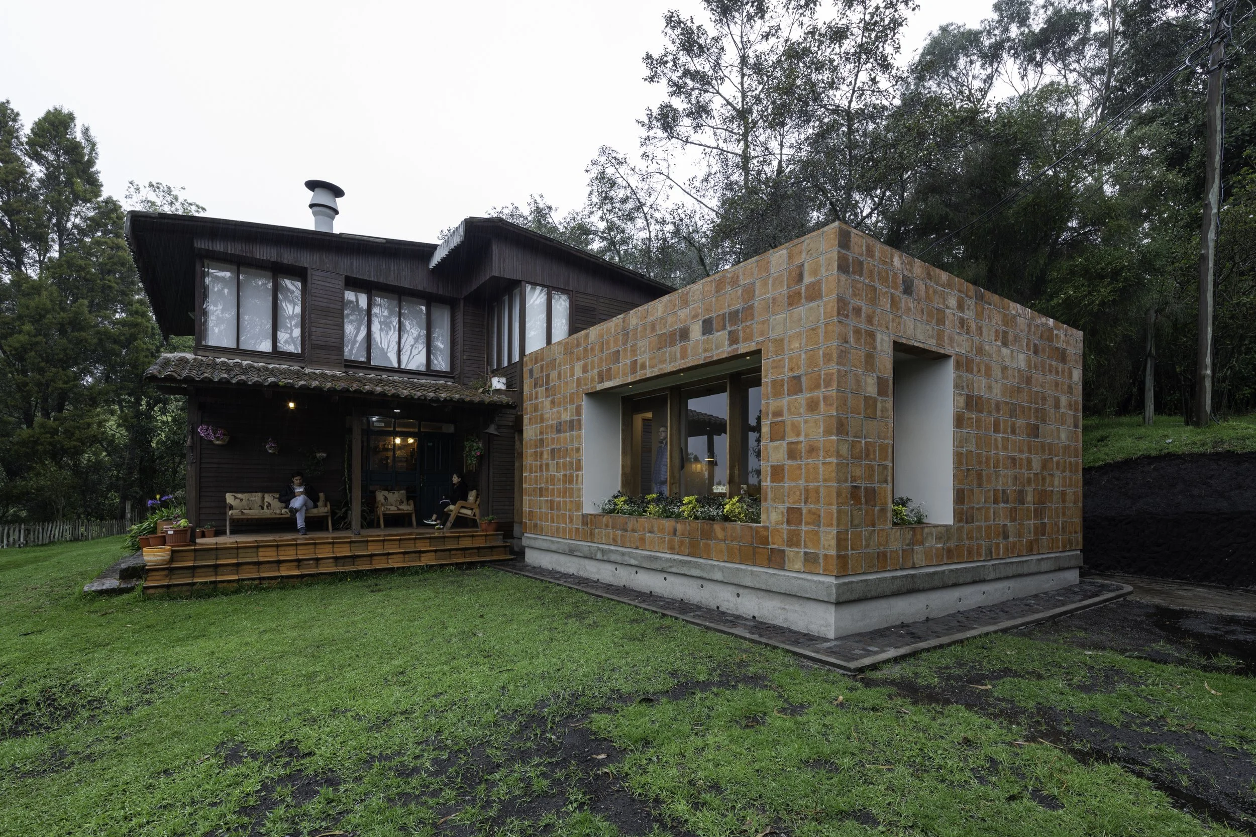 Casa moderna con estructura de ladrillo y madera, rodeada de césped verde y árboles, con personas sentadas en el porche.