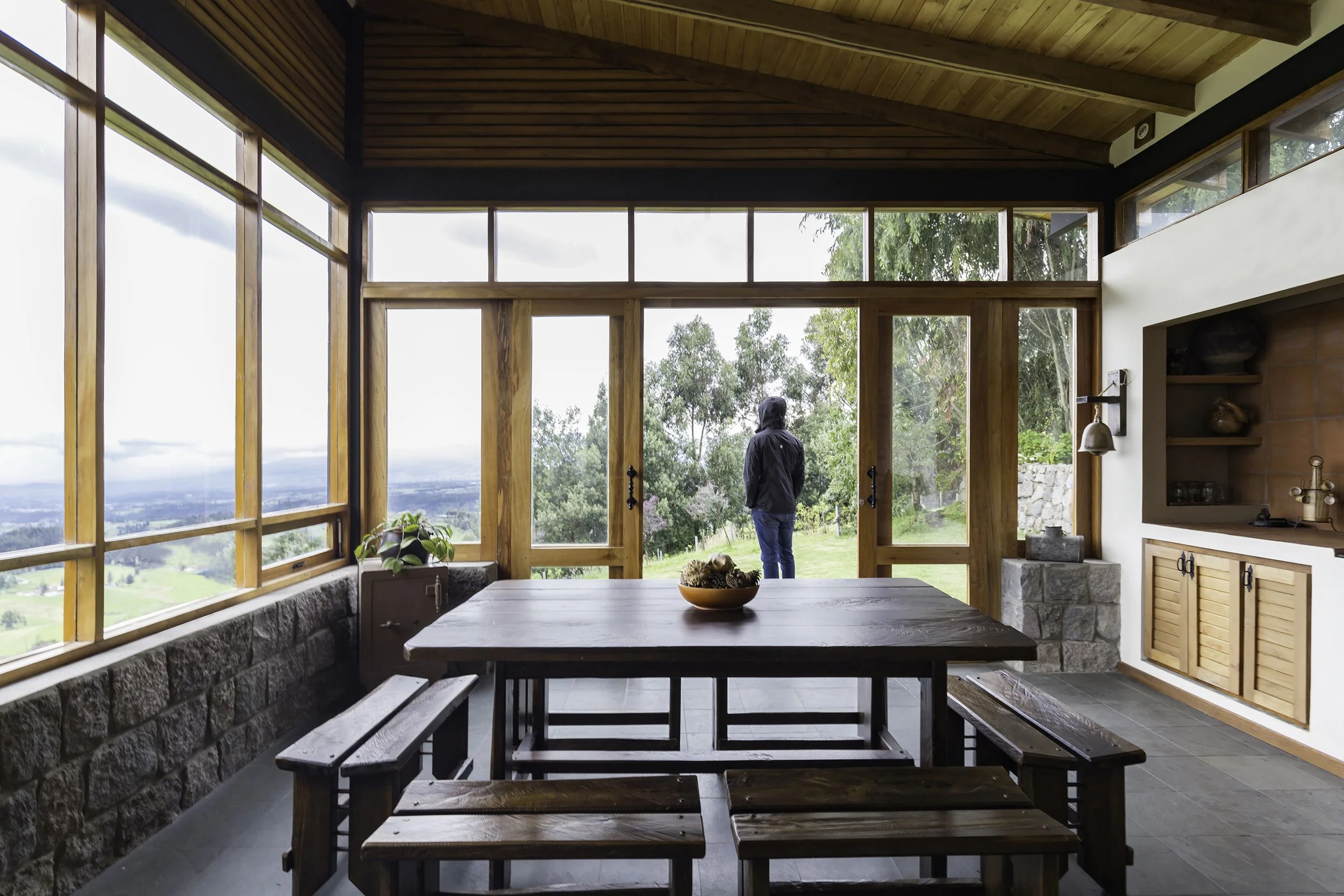 Interior de una casa de madera con ventanas grandes, vista hacia un patio con árboles, una persona de espaldas y un perro pequeño.