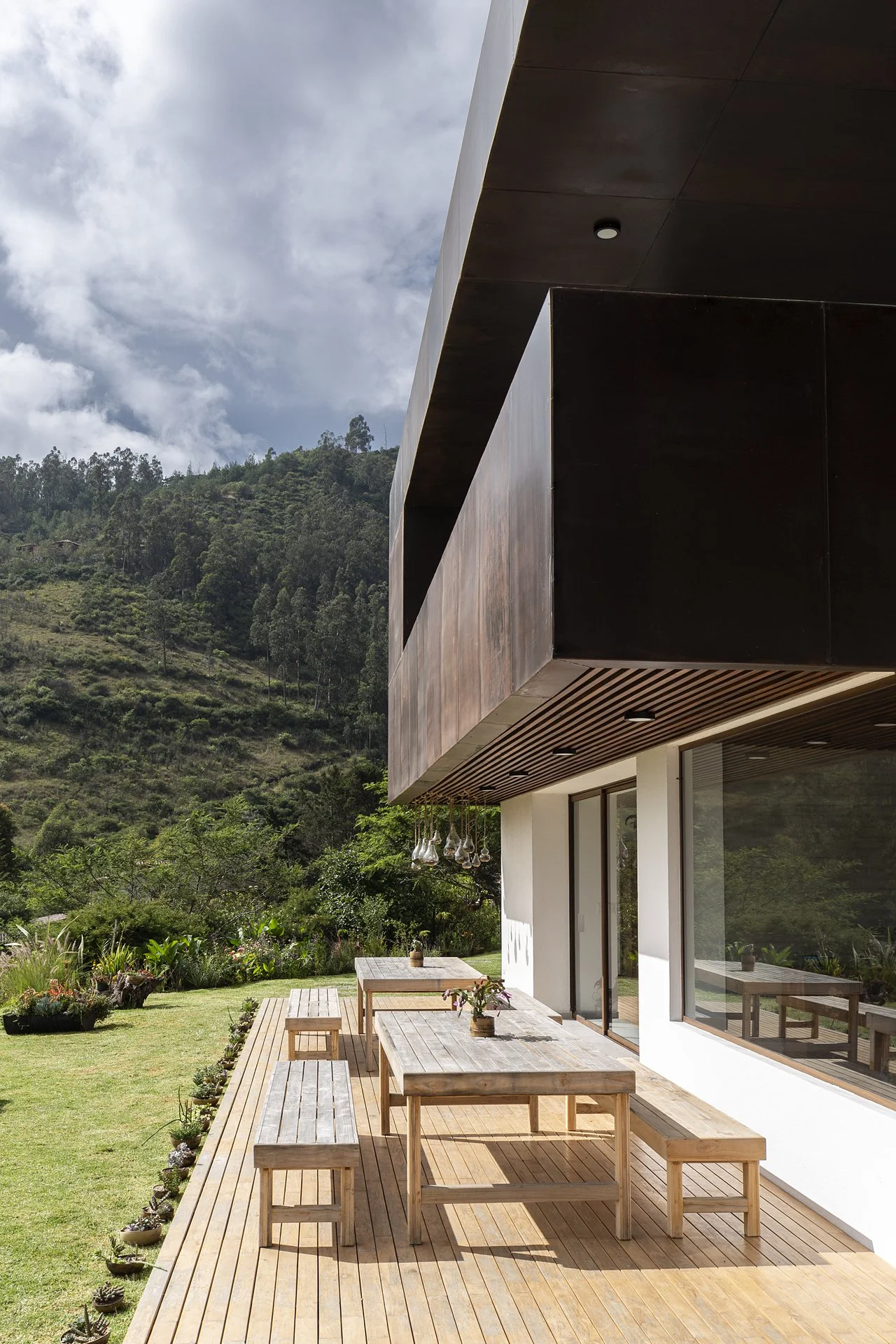 Vista de una casa moderna con balcón de madera, mesas y bancas en un jardín, con árboles y montaña al fondo, bajo cielo nublado.