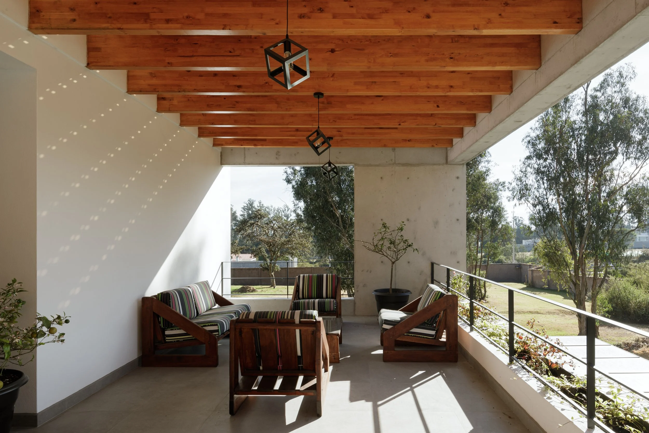 Espacio al aire libre techado con madera y concreto, con mobiliario de madera y cojines con rayas de colores, plantas, vista a árboles y cielo