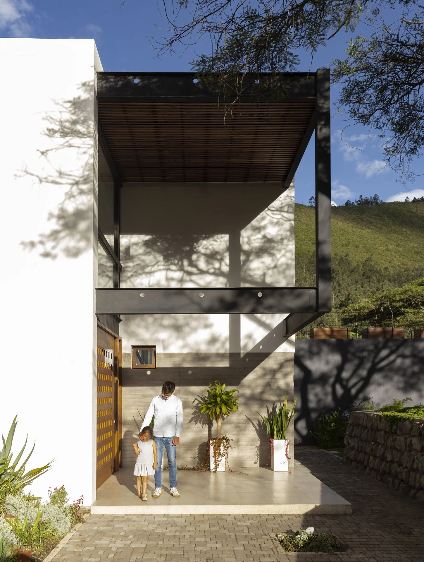 Una casa moderna con balcón y una entrada con dos personas, un adulto y una niña, caminando en el patio delantero, rodeados de plantas y bajo un cielo despejado.