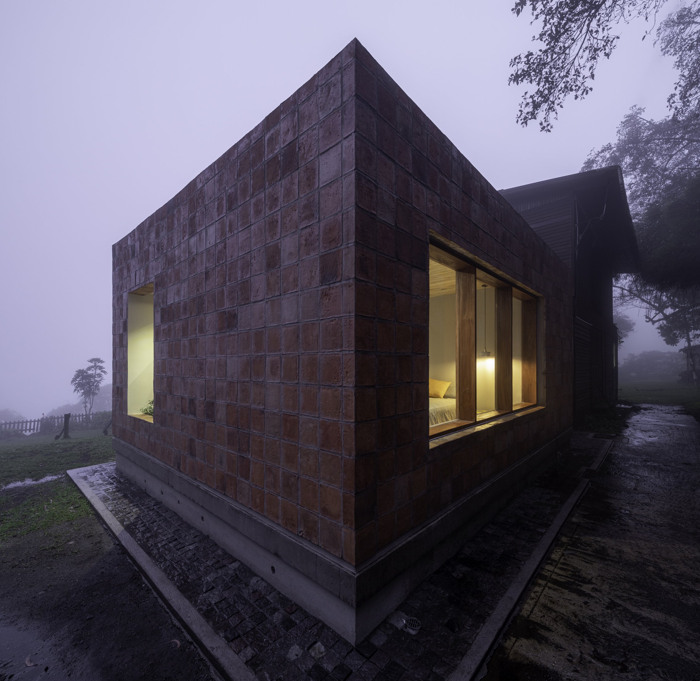 Edificio de ladrillos rojos con ventanas de madera y luz interior, en un entorno con niebla y árboles en el fondo.