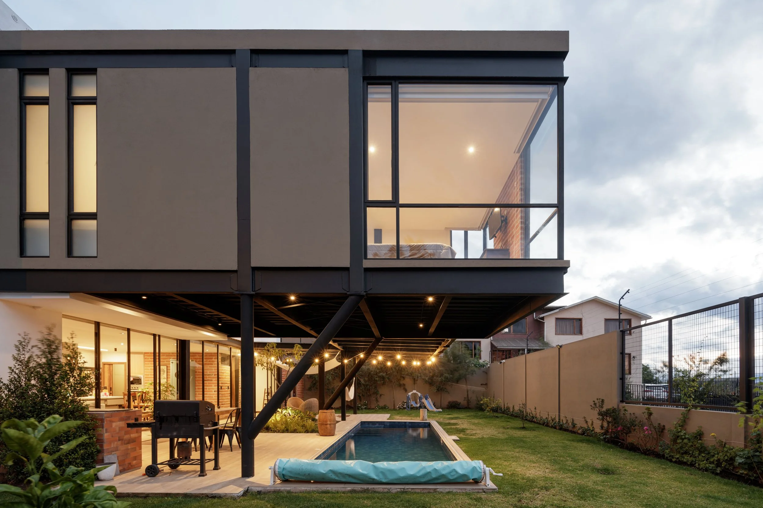 Casa moderna de varios niveles con patio y piscina, vista desde el jardín, con luces exteriores encendidas en la noche.