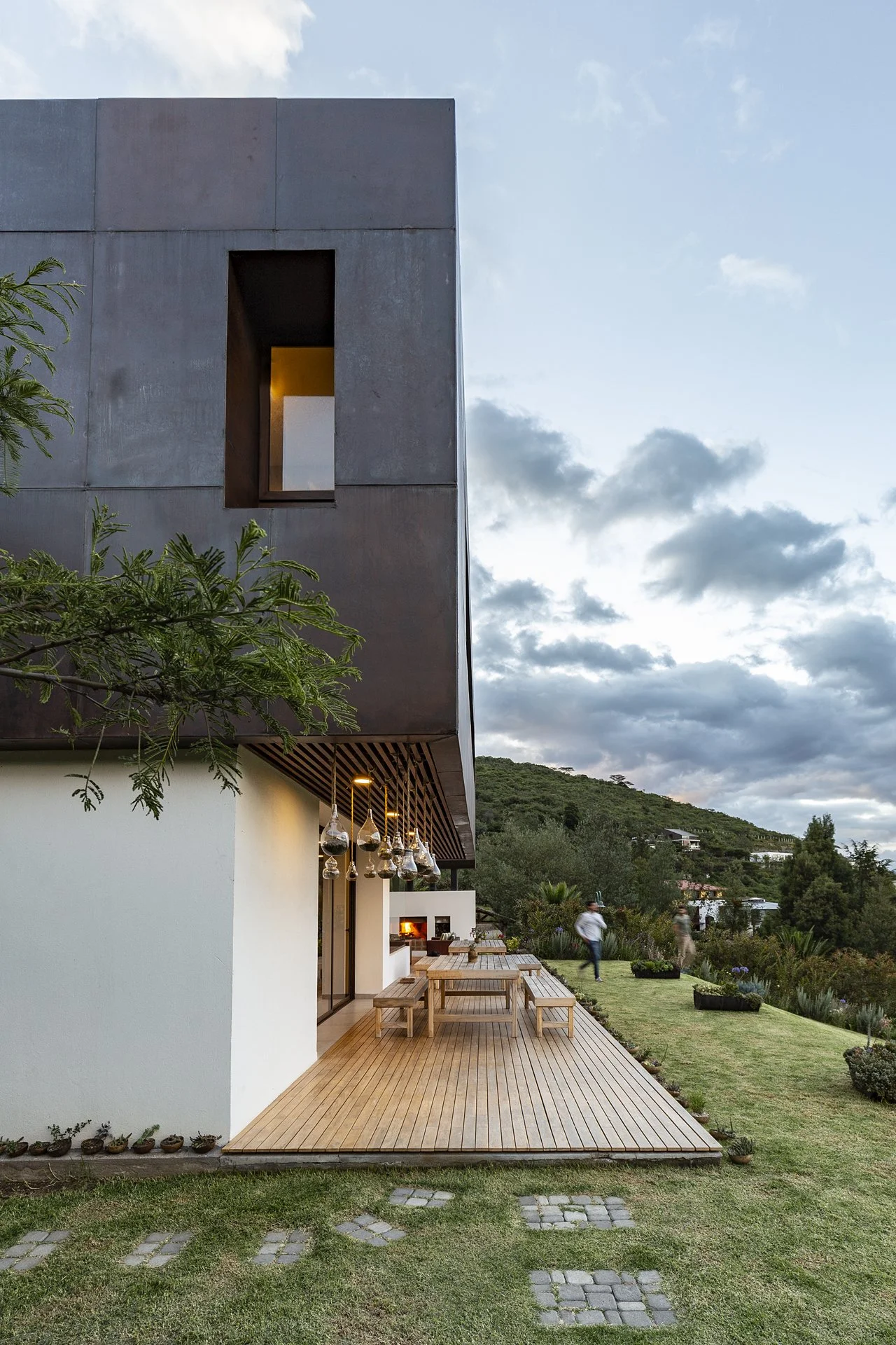 Casa moderna con terraza de madera, áreas verdes y vista a las montañas, en la tarde con cielo parcialmente nublado.