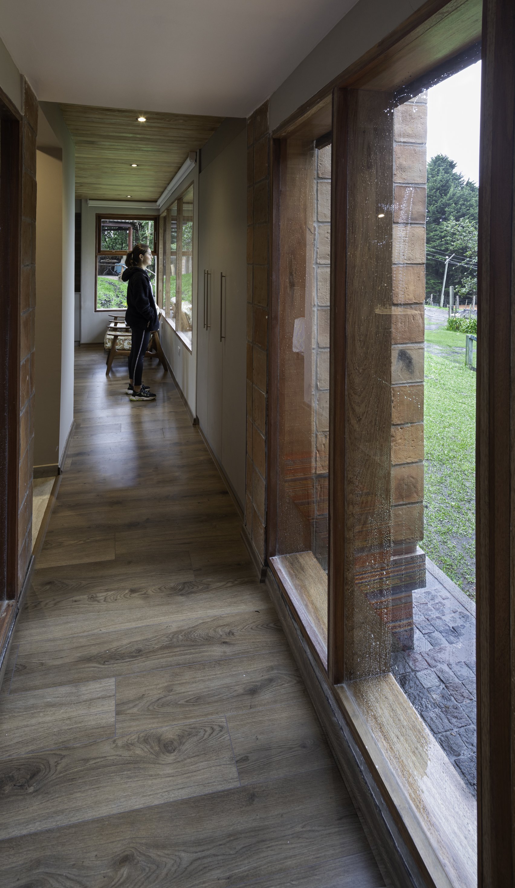 Vista de un pasillo interior con paredes de ladrillo y ventanas grandes, por donde se ve un jardín con árboles y césped; una persona observa por la ventana.